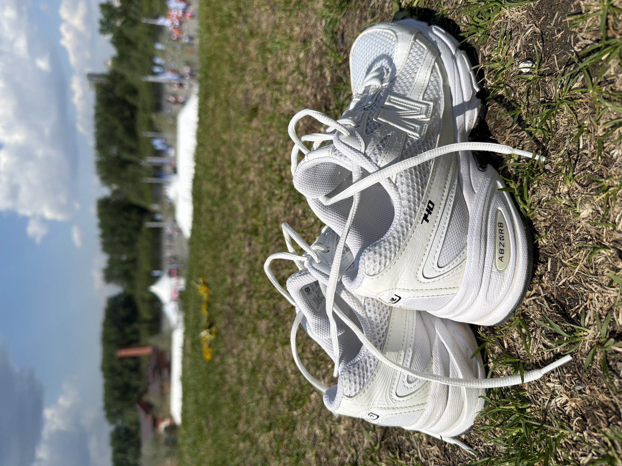 A pair of white New Balance 740 running shoes sit on a patch of green grass. The shoes are side-by-side, with their laces tied loosely. In the blurred background, there are trees, a white tent, and what appear to be people gathered, suggesting an event is taking place. It is not possible to confirm the location in Tyumen, Russia based on the visual data provided.