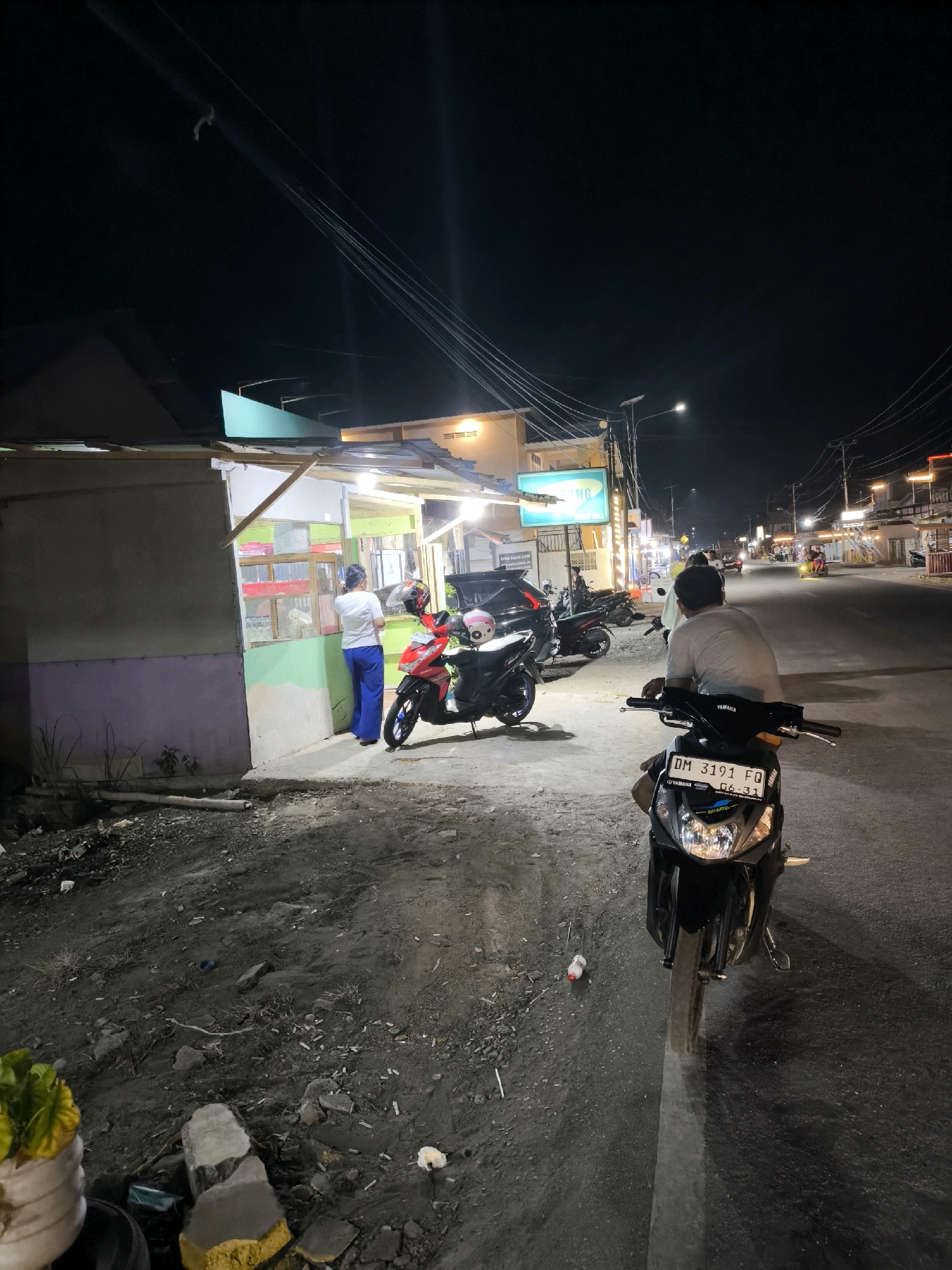 The image depicts a dimly lit street scene at night, possibly in Moluo, Indonesia. On the left, a small, open-fronted shop or stall is visible. A person wearing a white shirt and blue pants stands near the entrance of the shop, facing away from the camera. In front of the shop, a red motorcycle is parked. In the foreground, another motorcycle, black with a visible license plate reading "DM 3191 FQ", is positioned facing the street, with its headlights on. A person wearing a grey t-shirt is seated on this motorcycle, their back to the camera. The street itself is paved and appears to be a road with traffic lights and streetlights visible in the distance. Other motorcycles and a few people can be seen further down the street. The ground on the left side of the image is unpaved and appears to be dirt or gravel, with some debris scattered around.