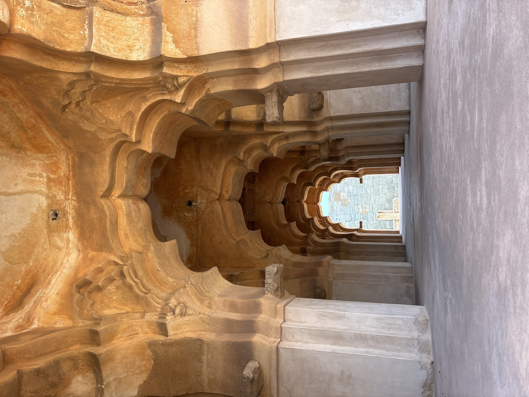 A ground-level, interior shot depicts a deep, diminishing corridor, angled slightly upward toward a distant opening revealing an exterior stone structure. The corridor's left side features an elaborate, ornate wall with repeating, scalloped archways in a warm, earthy tone. These archways are stacked in layers, creating a sense of depth and intricacy. The wall transitions to a cooler, pale tone further into the corridor. The right side of the corridor has a smooth, lighter-colored wall with horizontal lines suggesting steps or tiers leading upward. Overhead, a series of simple, rectangular beams span the corridor, providing a structural framework. The lighting is uneven, with the foreground walls being more intensely lit than the darker, recessed areas. In the distance, the opening to the exterior reveals a section of stone construction. The image is from Hampi, Kamalapura, India.