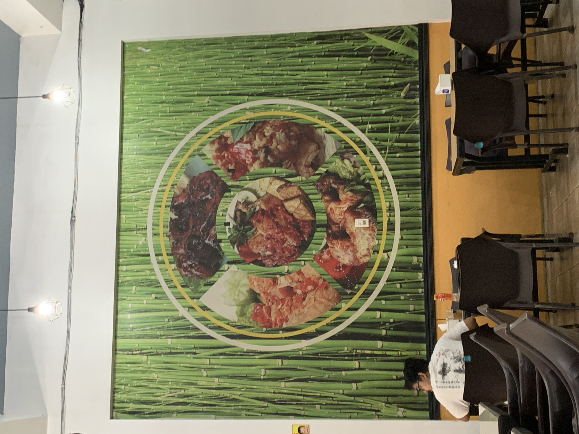 An indoor scene at a restaurant depicts a large mural of a bamboo forest with a circular arrangement of meat dishes in the foreground. A person wearing a white t-shirt and glasses sits at a table facing the mural, with their back to the camera. Several dark-colored chairs and tables are visible, along with a bottle of drink on a table. The walls are white, and a yellow accent color is present on the lower wall. The lighting fixtures appear to be industrial-style hanging lamps. The restaurant is located in Palohlada, Indonesia.