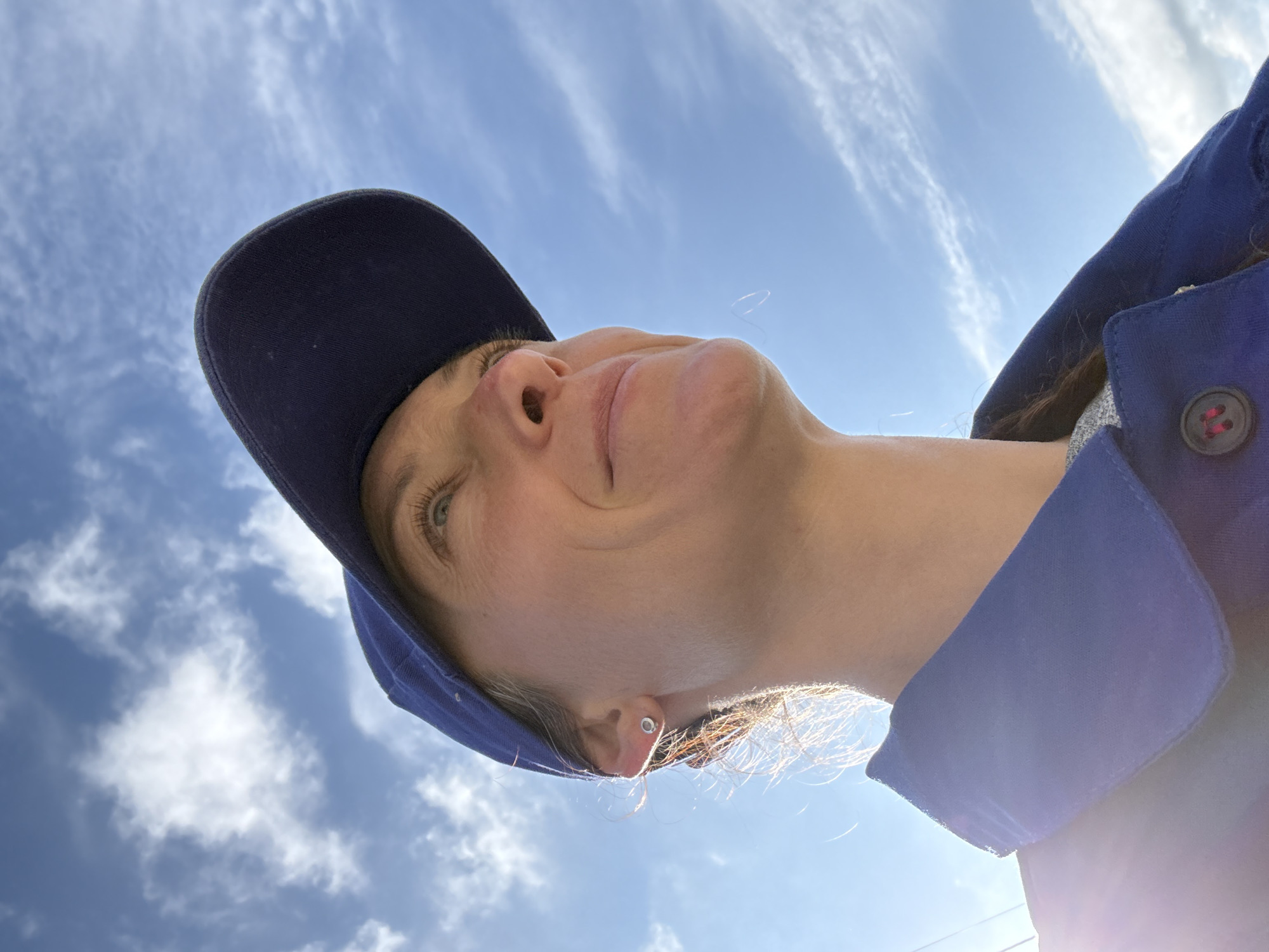 A low-angle shot shows a person with light skin looking up at the sky. The person is wearing a dark blue baseball cap and a blue button-down shirt. Wisps of blonde hair are visible near the ear. The sky is blue with scattered white clouds. The photo appears to be taken outdoors on a sunny day. Location is undetermined based on the image.