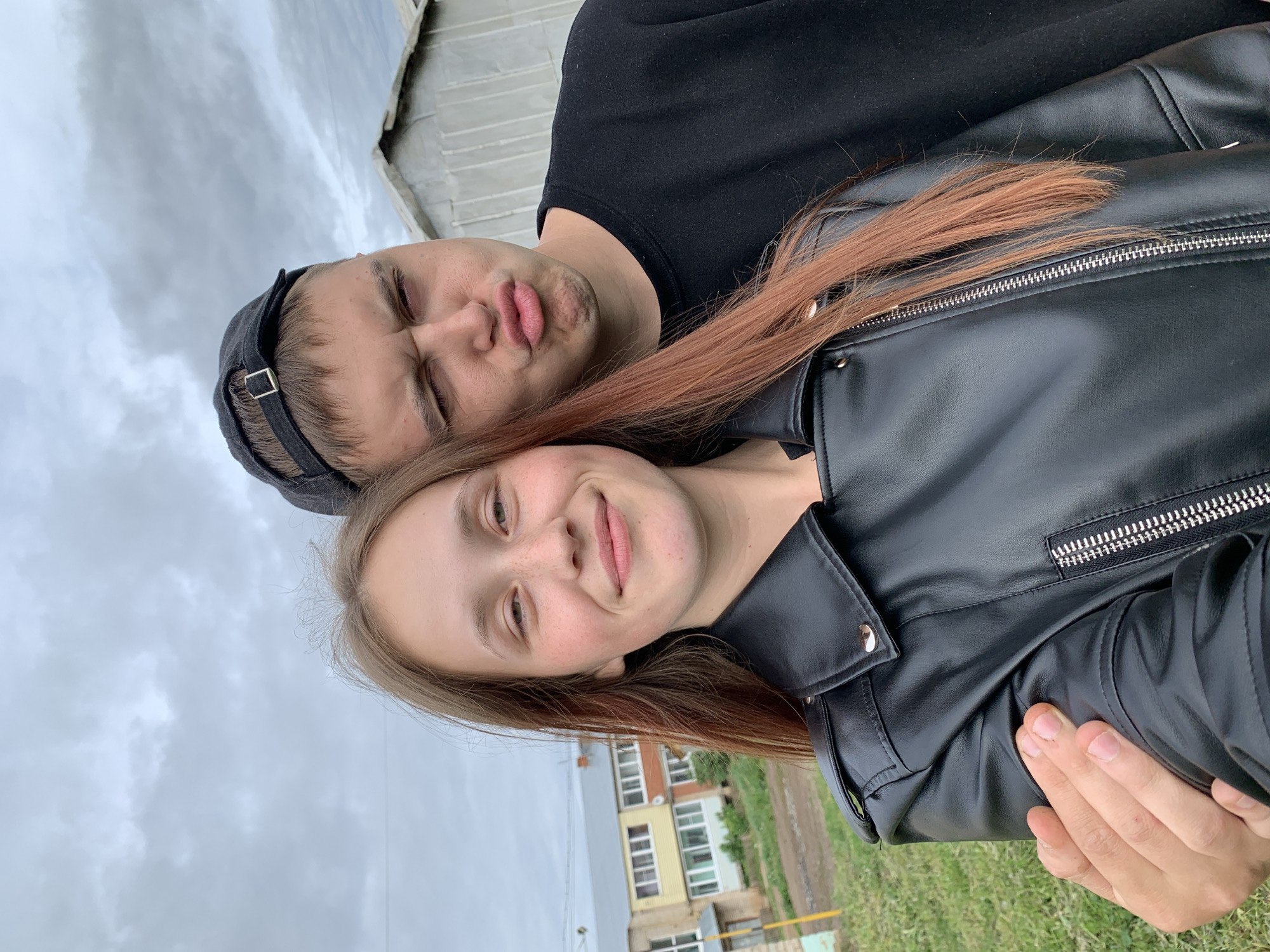 Two individuals pose for a selfie outdoors. A woman in a black leather jacket smiles, while a man behind her, wearing a black cap and black shirt, puckers his lips. The background includes a building and cloudy sky. The location is Фоки, Russia.