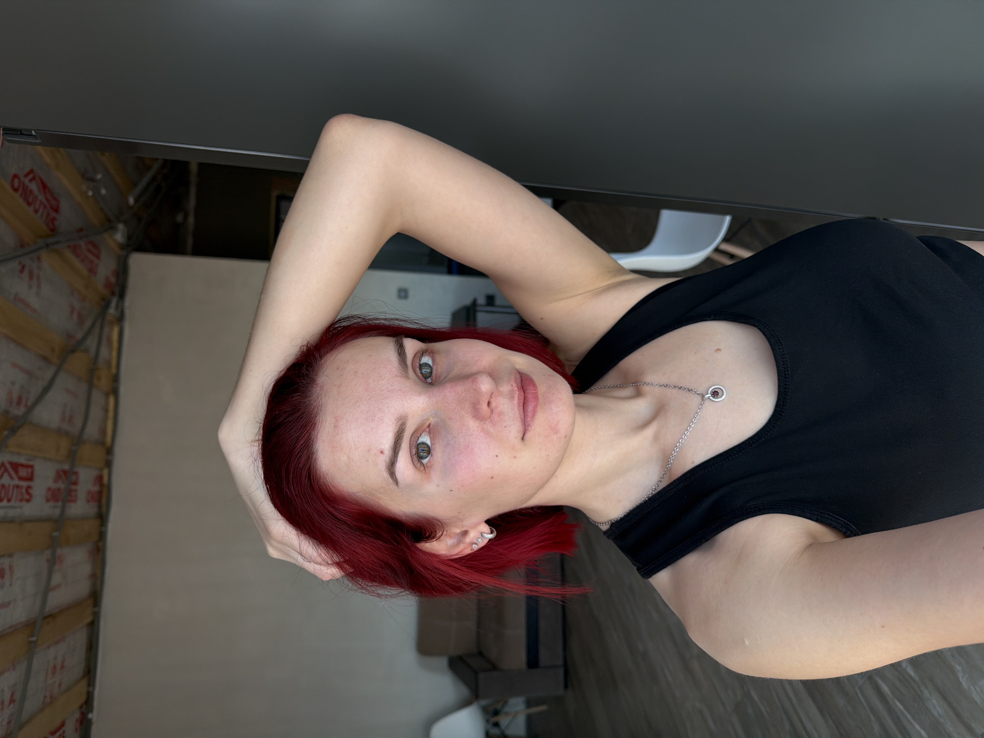 A young woman with red hair and a black top is posing for a selfie indoors. She has fair skin, freckles, and green eyes. Her right arm is raised above her head, and she is looking directly at the camera with a slight smile. She is wearing a silver necklace. The background includes a wall with exposed building materials and a glimpse of a room with a white chair and dark furniture. The location is Chaikovsky, Russia.