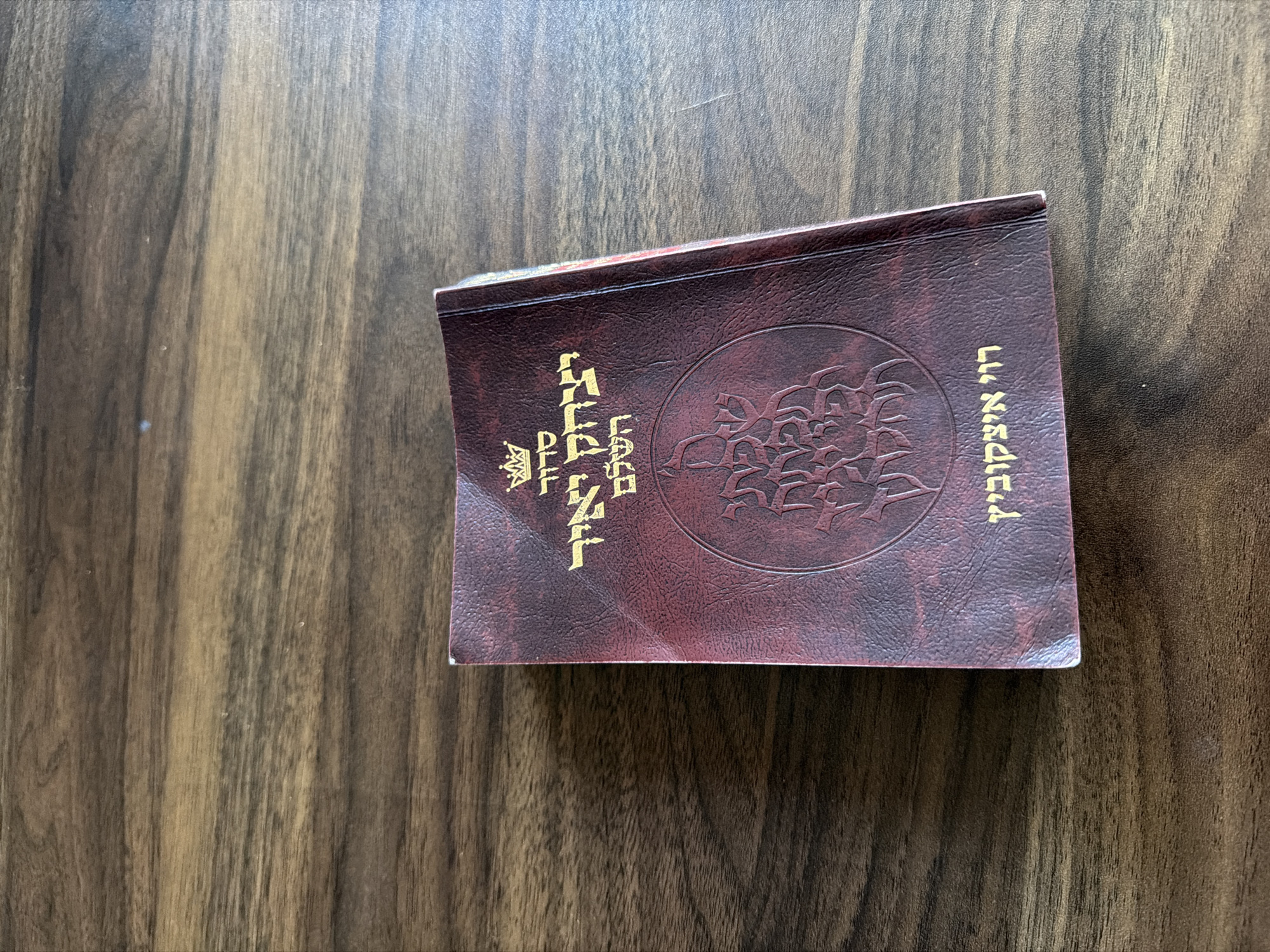A dark red, textured book with gold Hebrew lettering and an embossed design lies diagonally on a wooden surface. The book's cover, possibly faux leather, shows slight wear, particularly at the corners.
On the left side of the book, printed vertically in gold, is the Hebrew text "תָּהֳרַת הַמִּשְׁפָּחָה" (Tohorat HaMishpacha), which translates to "Family Purity." Below this, also in gold, is "הלכה למעשה" (Halakha Lema'aseh), meaning "Practical Law" or "Applied Jewish Law." A small, golden emblem, possibly a decorative crown or logo, is situated above the lower line of text.
On the right side of the book, also oriented vertically in gold, is the Hebrew phrase "זכר מנחם" (Zecher Menachem), meaning "In Memory of Menachem." In the center of the cover is a large, circular embossed pattern with intricate floral or abstract designs.
The background is a wooden surface with a visible grain, featuring warm brown tones with darker and lighter streaks. The lighting is soft and even, suggesting an indoor setting. No activity is taking place; the image is a static shot of the book.