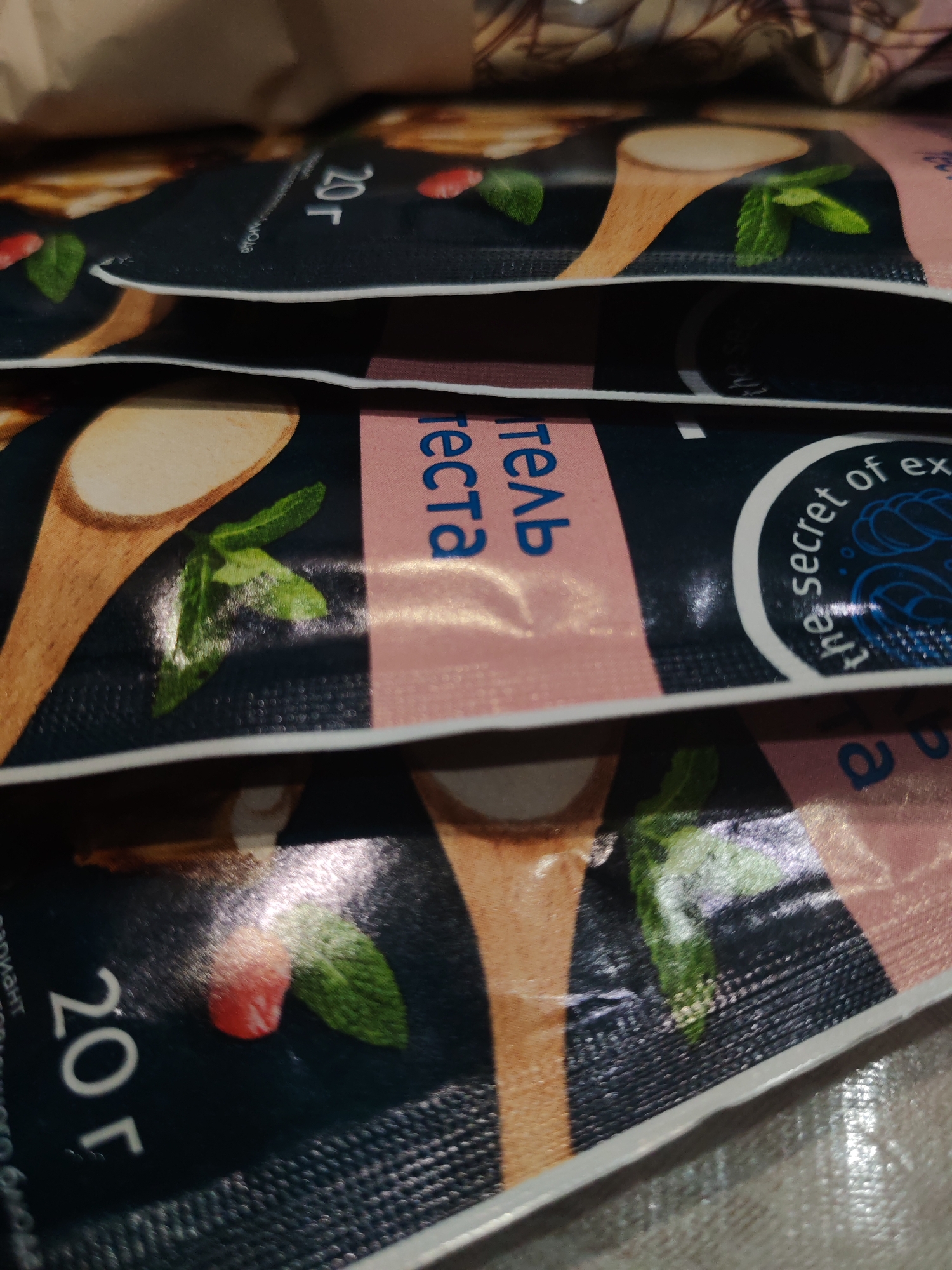 Here is a close-up shot of several small, rectangular sachets stacked on top of each other. The sachets are predominantly dark, possibly black or dark blue, with a pinkish-peach colored section along the right side. Each sachet features an image of a wooden spoon with a white substance, along with some green leaves that appear to be mint. There is a red fruit, possibly a raspberry, on each packet. Text in Russian is visible on the pink section of the sachets, reading "гель теста". A circular design with the text "the secret of ex" is also present. The sachets are labeled with "20 г". The background is out of focus, but appears to be a plain surface.