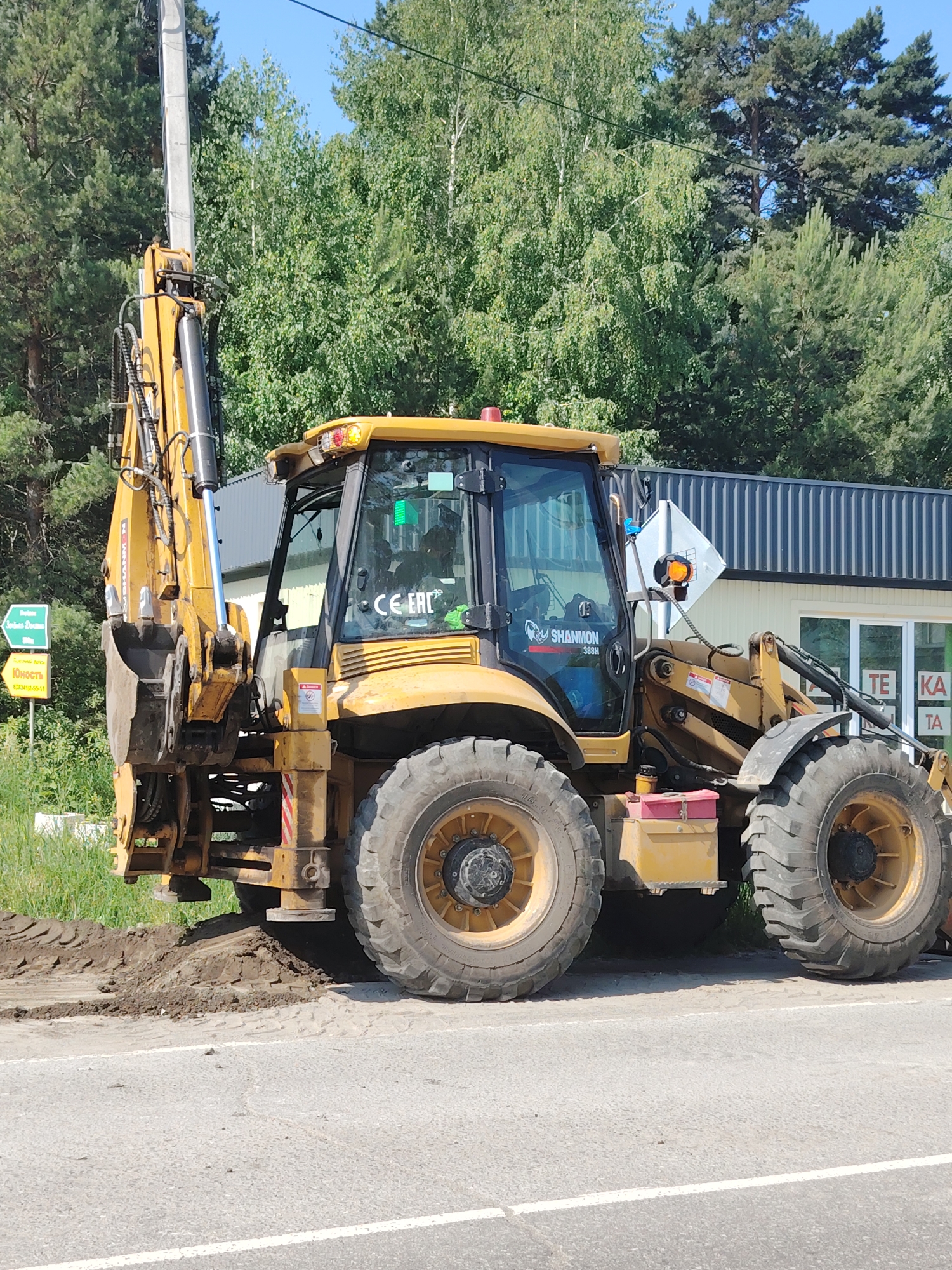 An outdoor, daytime, medium shot captures a yellow backhoe loader parked on the side of a paved road. The vehicle's front loader is positioned low, and its rear digging arm is raised and extended. The backhoe has a cab with tinted windows, one of which displays a green sticker. A red toolbox is visible beneath the cab, and a yellow storage compartment is mounted on the side. Two large tires are prominent on the right side of the vehicle, and a portion of the front loader's tires are visible on the left. The ground in front of the backhoe is disturbed earth, with tire tracks visible. Behind the backhoe, a row of trees forms a dense green backdrop against a clear blue sky. To the left of the backhoe, a white utility pole stands tall, and a green directional sign with yellow text is mounted on it. The background also includes a building with a corrugated metal roof and windows, and a white diamond-shaped sign with orange and black details is mounted on a pole near the backhoe. The overall scene suggests construction or road work in progress.