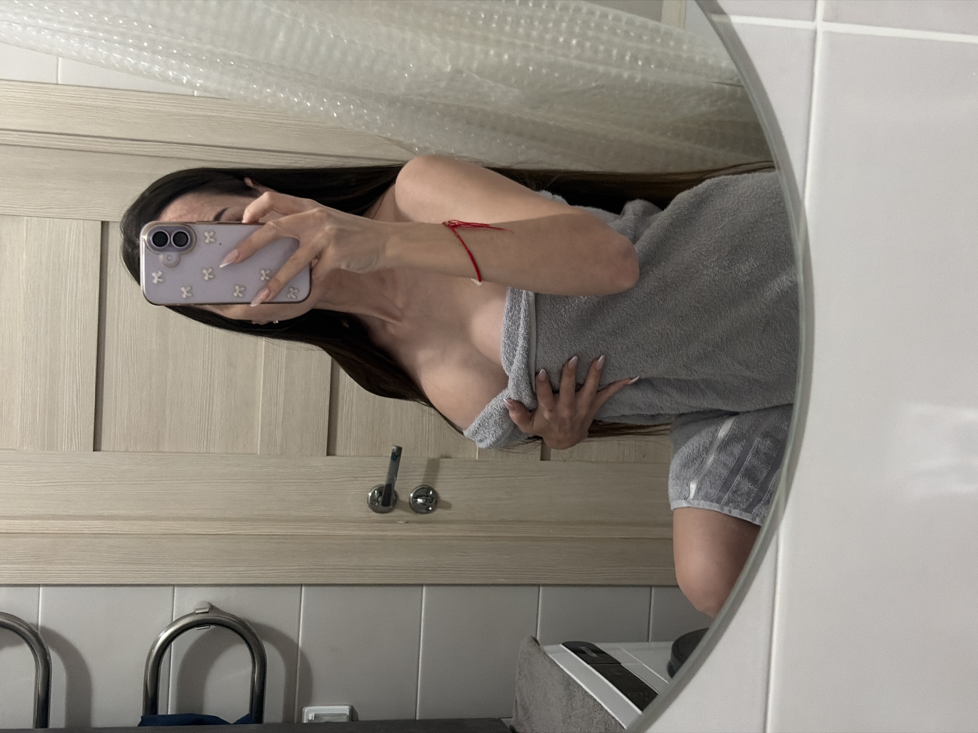 The image captures a mirror selfie of a young woman in a bathroom, presented with a 90-degree counter-clockwise rotation. The woman, with long dark hair, is wrapped in a gray towel that covers her chest and extends to her upper thighs. A thin red string bracelet is visible on her left wrist.
She holds a light purple smartphone, equipped with a dual-lens camera, which has a case decorated with small white flower-shaped patterns. Her manicured nails are long and light-colored, featuring white tips. Her face is mostly obscured by the phone as she takes the photo.
The background, reflected in the mirror, consists of light wooden wall panels, likely part of a vanity or cabinet, above sections of white and light gray tiles. Metallic faucet handles or towel rings are visible below the wooden paneling. The upper part of the mirror reflects a distorted view of clear, possibly bubble-wrap, material. While not depicted in the image, the provided context indicates the location is Saint Petersburg, Russia. No visible text is present in the image.