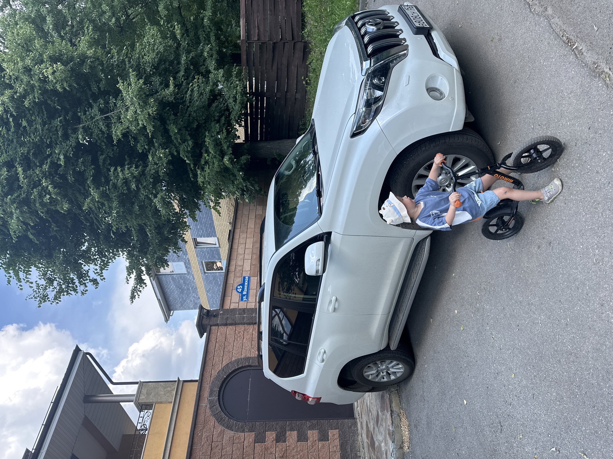 In Nizhny Novgorod, Russia, a young boy wearing a white bucket hat, a blue t-shirt, and denim shorts rides a black balance bike on an asphalt street. He is positioned to the right of a white Toyota Land Cruiser Prado. The car is parked next to a brick wall, which has a dark arched gate. Behind the car and wall, a wooden fence is visible, bordering a grassy area with trees. A blue and yellow multi-story building with windows is visible in the background, along with another building with a tan facade and a balcony. A street sign with the number "45" and Russian text "ул. Манежная" (Manézhnaya Street) is attached to the brick wall. The sky is partly cloudy.