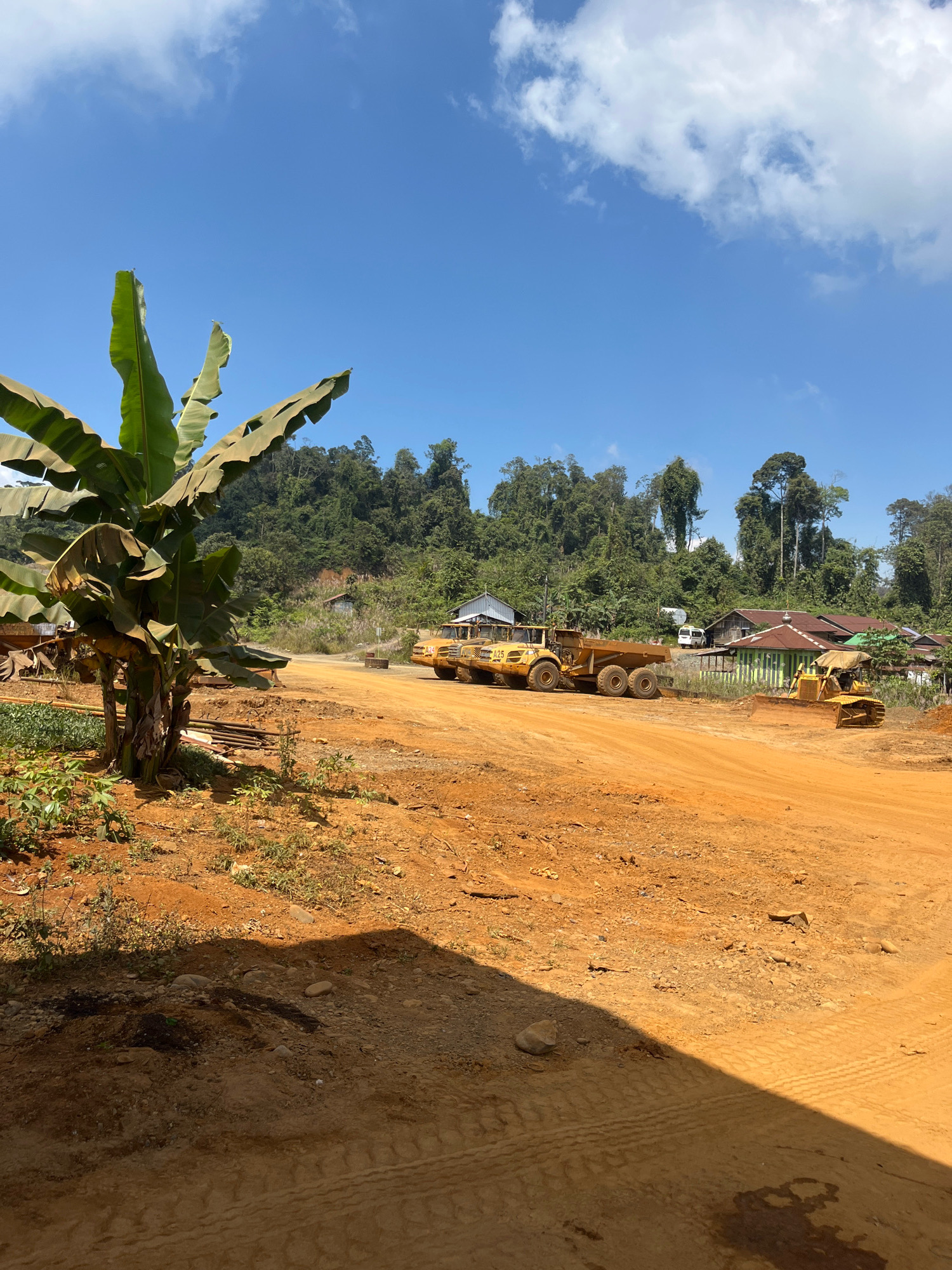 The image depicts an outdoor construction site under a clear blue sky with scattered clouds. In the foreground, a large banana plant with broad leaves stands to the left. The ground is covered in reddish-orange dirt, with visible tire tracks and some scattered rocks and vegetation.
Further into the scene, several yellow construction vehicles are present. On the left, a large articulated dump truck is parked. To its right, a bulldozer is actively working, pushing dirt. In the background, a small village or settlement is visible, with several buildings and a white van. The village is nestled against a densely forested hillside. The overall impression is of a rural area undergoing development or resource extraction. The specific location is stated as Unknown, Indonesia.