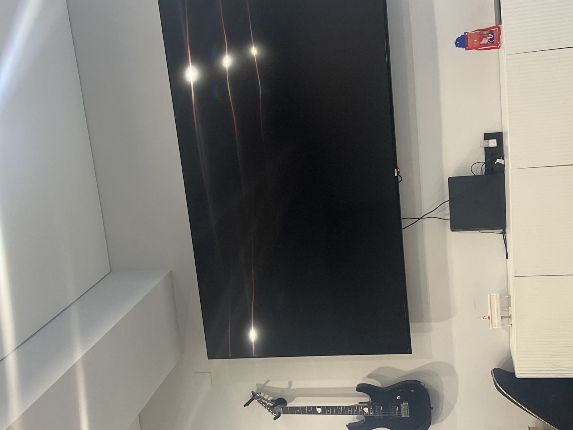 The image depicts an indoor setting, likely a living room or entertainment area, with a large, black television screen dominating the scene. The screen displays reflections of light, suggesting ambient lighting in the room. To the right of the television, a white, ribbed cabinet is visible, along with a black gaming console mounted on the wall below it. A red and blue toy, possibly a water gun, is placed on top of the cabinet. Below the television, an electric guitar with a black finish is displayed horizontally, mounted on the wall. The overall atmosphere is casual and suggests a space for entertainment. The lighting appears to be artificial, and there are no explicit indicators of the time of day or weather conditions. No text is visible in the image.