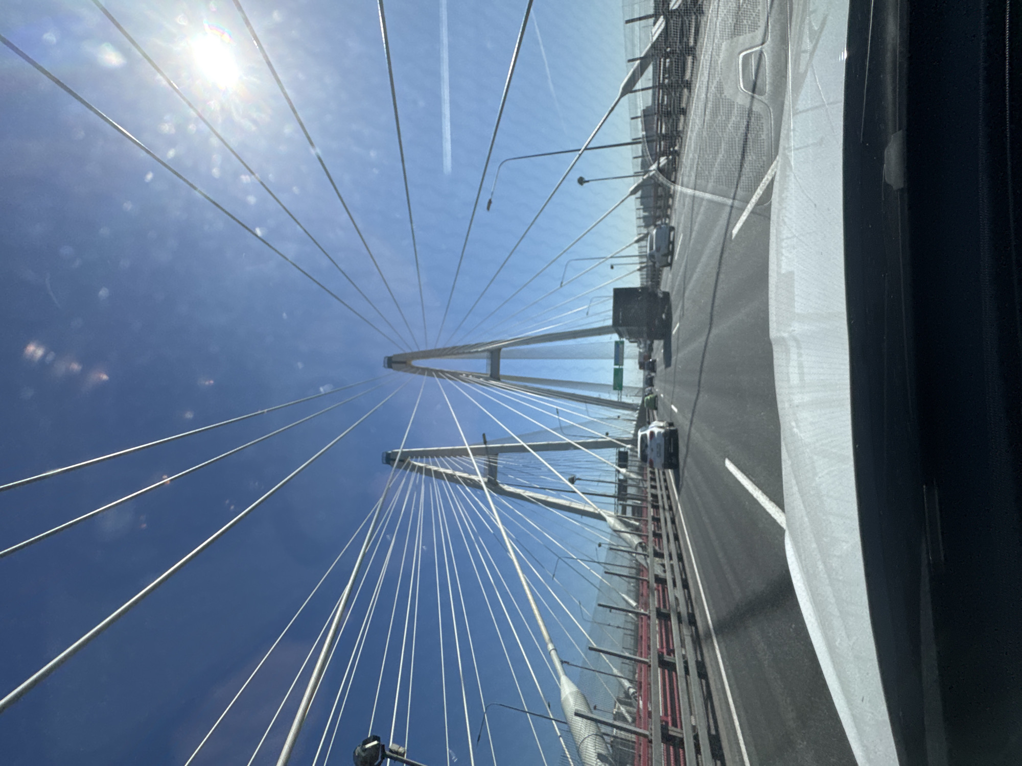 The image is a wide-angle, forward-facing view from inside a vehicle traveling on the cable-stayed Big Obukhovsky Bridge in Saint Petersburg, Russia. The bridge's distinctive twin towers and numerous white cables dominate the sky, which is bright blue with the sun shining strongly, causing lens flare and reflections on the windshield. Traffic is visible on the multi-lane highway, including a white SUV in the right lane and a dark truck in the left lane. A white car is also visible in the distance on the left. The bridge structure, including railings and lighting poles, is present along the sides of the road. The foreground shows the hood and dashboard of the vehicle from which the photo was taken.