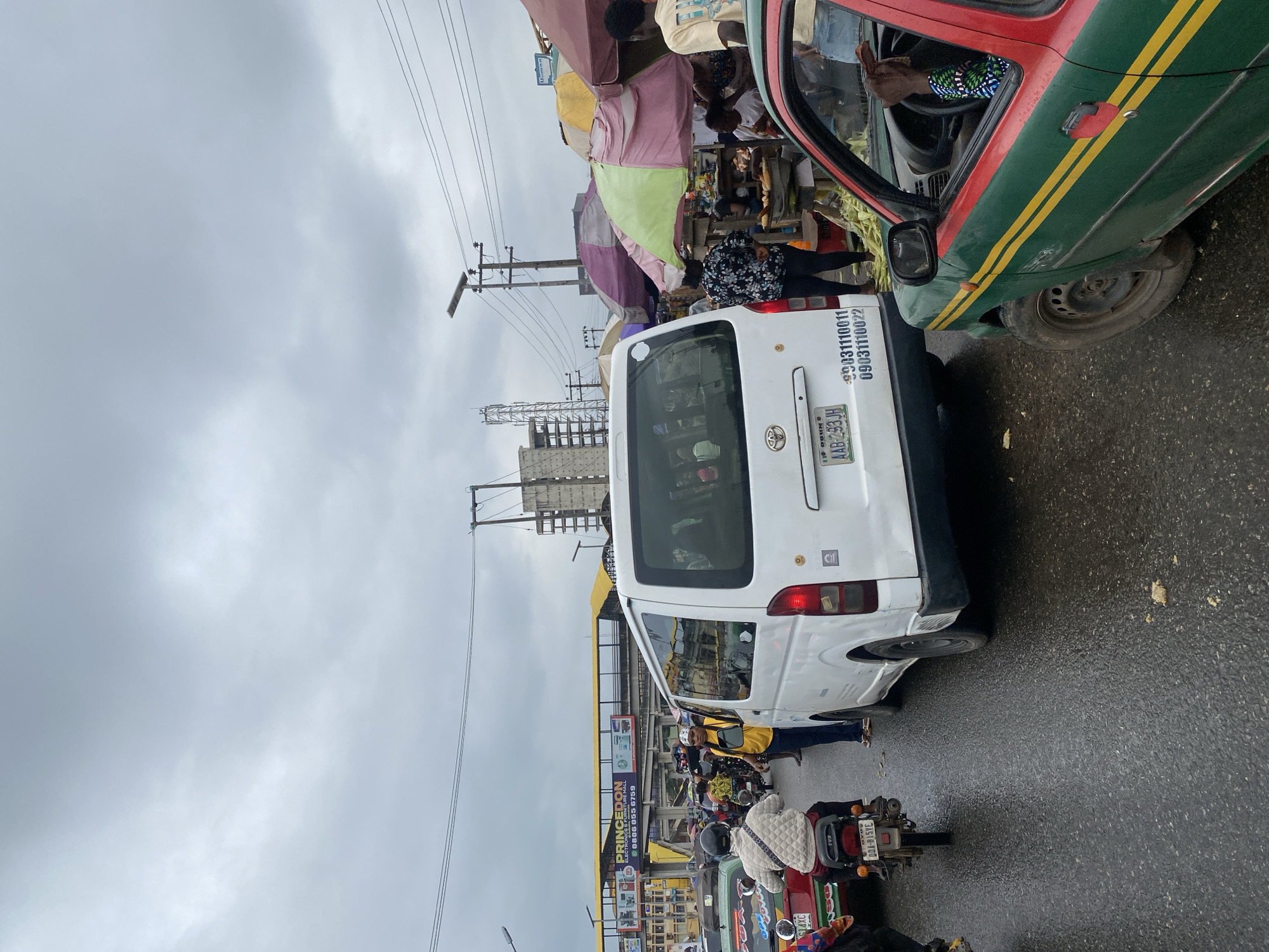 The image depicts a busy street scene in Abeokuta, Nigeria, under an overcast sky. In the foreground, a white van with the license plate AAB-293JH is stopped in traffic. To the left of the van, a motorcyclist wearing a patterned jacket is visible. To the right, a green and red taxi is partially visible, with the driver's arm resting on the open window.

Behind the van, a street market is set up with various stalls under colorful umbrellas. People are seen interacting at the market, some appearing to be vendors and others shoppers. Several motorcycles and other vehicles are present, indicating a congested traffic situation.

In the background, a yellow pedestrian bridge with signage for "PRINCEDON ELECTRONICS & FURNITURE MALL" spans across the road. Power lines and utility poles are visible overhead. Construction on a building is also in the distance. The road appears wet, suggesting recent rain. The overall atmosphere is that of a typical bustling urban environment in Nigeria.