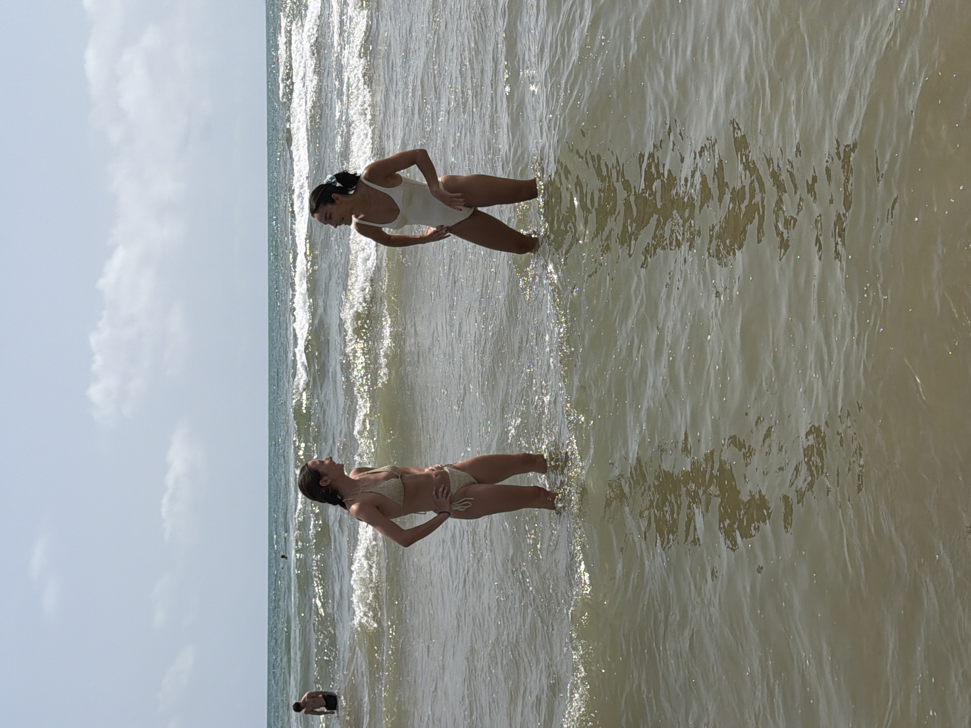 Two young women are standing in the ocean, waist-deep in water. The woman on the left is wearing a tan bikini, and the woman on the right is wearing a white one-piece swimsuit. Both women are facing each other and appear to be talking, with their hands on their hips. The ocean is choppy with moderate waves, and the sky is partly cloudy. In the distance, a few other people can be seen in the water. The overall setting is a beach on a sunny day.