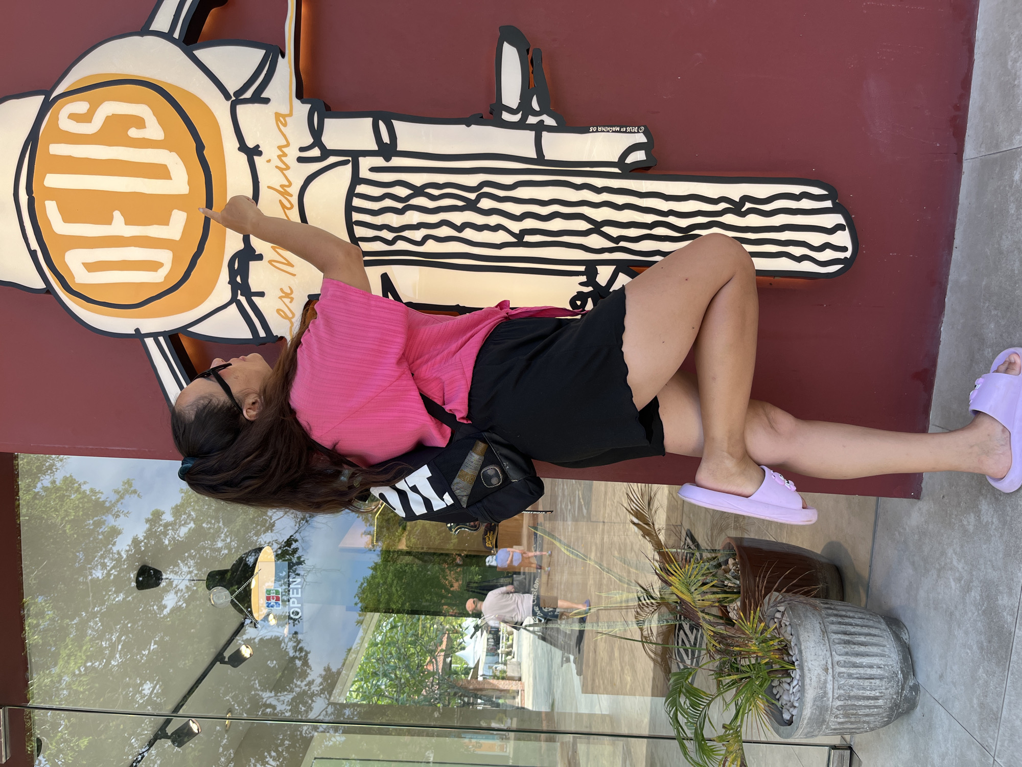 A woman with dark hair tied in a ponytail, wearing sunglasses, a pink top, and black shorts, stands with her right leg bent at the knee, pointing a finger at a large, stylized "DEUS" logo on a maroon wall. She is wearing light purple slides. A black backpack with white straps is on her shoulders. To her left, through a glass storefront, a man in a light-colored shirt and hat, and another person in blue, are visible walking outside. Two potted plants are situated to the left of the woman. The scene appears to be outdoors, near a building with a glass facade. The overall context suggests a street or commercial area in Kuta Selatan, Indonesia, likely near a Deus Ex Machina store given the logo.
