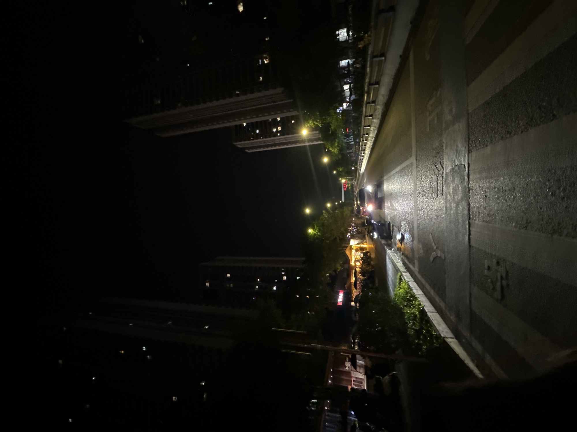 The image captures a nighttime scene on a street in the Chongqing High-tech Zone, China. The perspective is from a pedestrian crossing, looking down a multi-lane road flanked by tall buildings. The street is wet, suggesting recent rain, and illuminated by streetlights and the headlights of vehicles. On the left side of the road, there appears to be a strip of greenery and some low structures, possibly shops or food stalls, with people and umbrellas visible. Several cars are on the road, including one with its headlights on, approaching the viewer. The buildings on both sides of the road are tall and modern, with many windows showing interior lights. The overall atmosphere is that of a bustling urban environment at night, with the wet pavement reflecting the artificial lights.