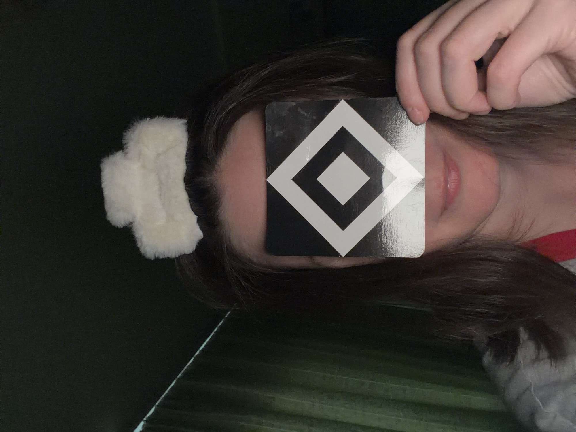 A person is holding a black and white geometric patterned card in front of their face, obscuring their eyes. The person has brown hair and is wearing a white furry accessory. The background is a dark green color. The location is Unknown, Unknown.