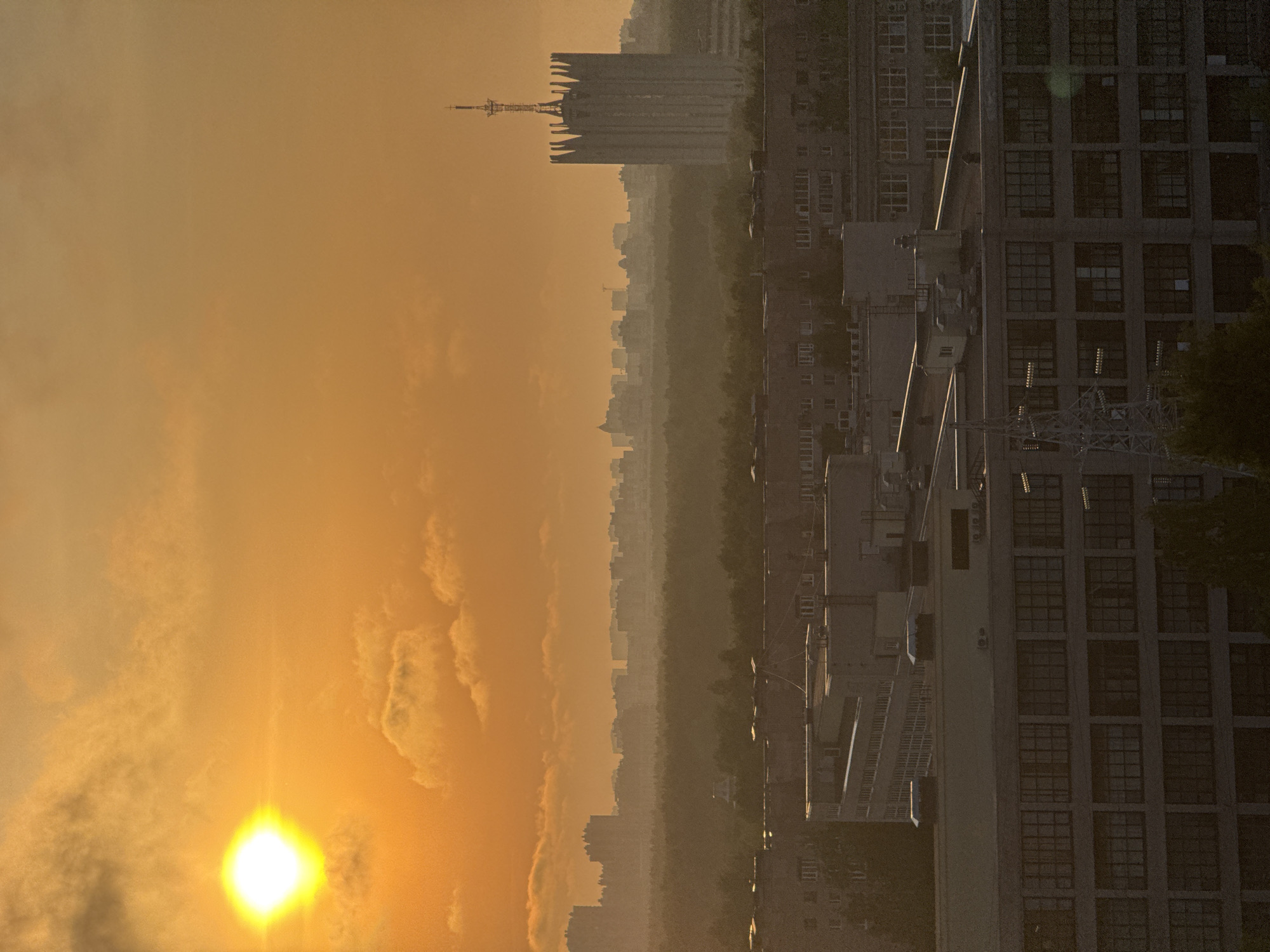 The image shows a cityscape at sunrise or sunset, likely in Sinyavino, Russia. The sky is a bright yellow-orange gradient, with the sun visible near the bottom left corner. The cityscape consists of a mix of low-rise and high-rise buildings, with a prominent tower with a communication antenna in the background. The buildings appear to be densely packed, with some greenery visible between them. The foreground features a large building with a grid-like window pattern, possibly a factory or residential complex. There is a stairwell and other architectural elements visible on the building's facade. The image has a hazy or slightly blurred appearance.