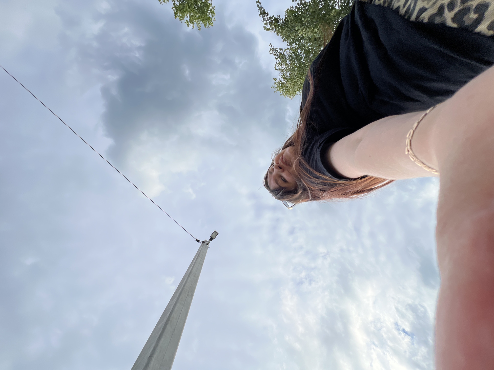 A low-angle, outdoor, wide shot captures a woman in a black t-shirt and leopard print skirt, looking up at the sky with a slight smile. She is wearing a delicate gold bracelet on her left wrist. Her right arm is extended towards the camera, with her thumb and forefinger obscuring part of the frame. Behind her, a grey, cloudy sky is visible, with a tall utility pole and a single wire stretching diagonally across the frame. Trees with green foliage are visible on the right side of the image. The scene is set outdoors, under a cloudy sky. The location is identified as Noviy, Russia.