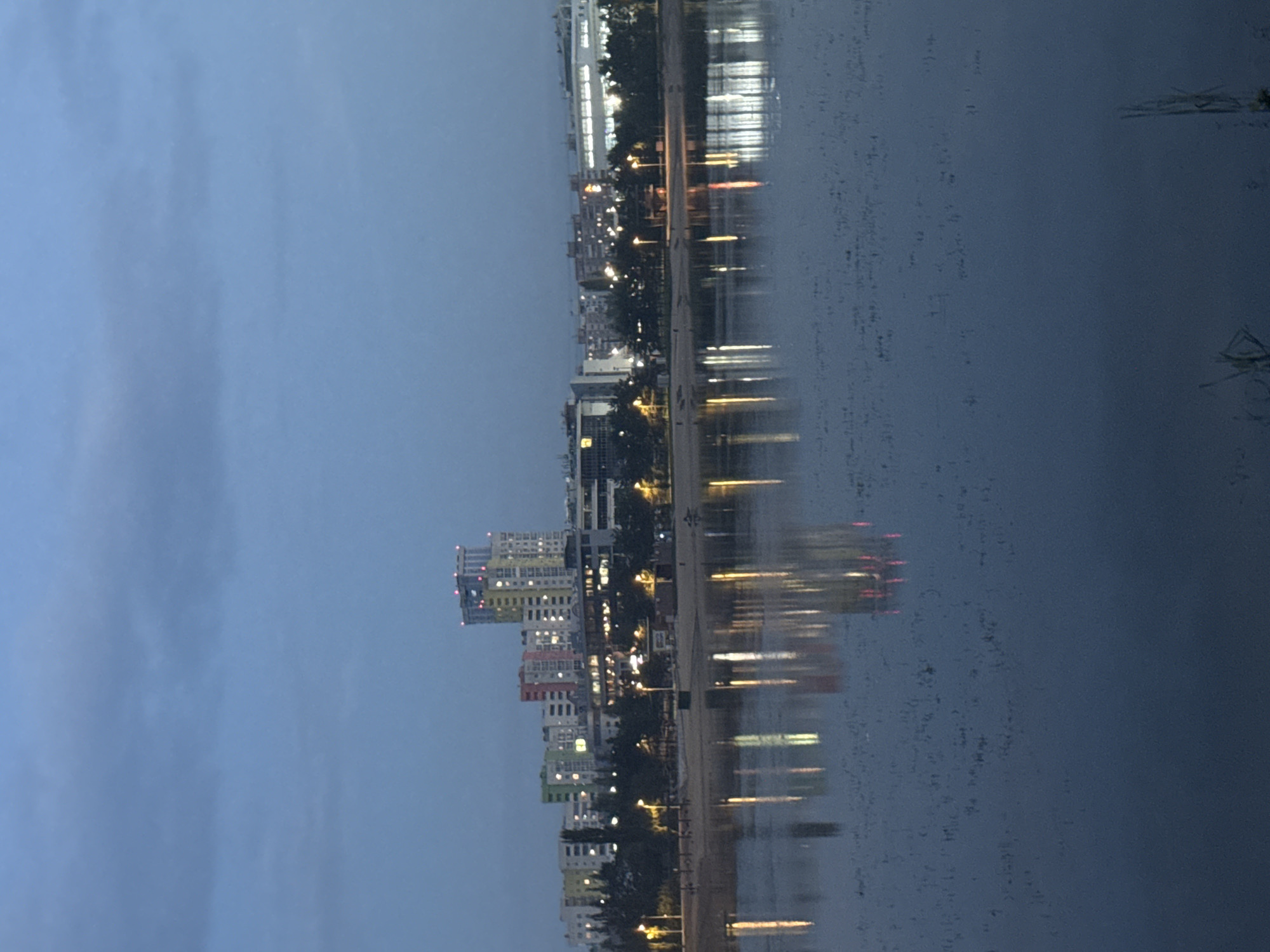 This is an outdoor, wide-angle, eye-level shot of a waterfront city skyline at dusk. The sky is a muted blue-gray with sparse clouds. In the mid-ground, a line of buildings stretches across the horizon. The buildings vary in height and architectural style, with some appearing to be apartment complexes and others more commercial. Many windows are illuminated, casting a warm glow. The most prominent structure is a tall, multi-story building with a light-colored facade and illuminated windows. A row of trees and streetlights line the area between the buildings and the water. The foreground is dominated by a body of water, likely a river or lake, which reflects the lights from the city, creating elongated, distorted streaks of color on the surface. The water is dark blue and shows subtle ripples. Some sparse vegetation is visible on the far right edge of the frame. There are no discernible people or animals in the image. The overall impression is one of a quiet evening in an urban setting. This appears to be a view of the Ufa city skyline, possibly along the Belaya River.