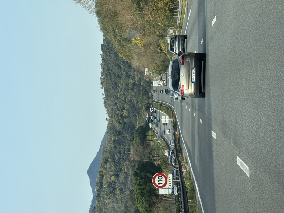 This image depicts a section of an active multi-lane highway, consistent with the A8 Autoroute du Soleil, located within or immediately adjacent to the city of Mandelieu-la-Napoule, France. The scene is observed from within a vehicle traveling forward.
The environment features a clear, light blue sky above rolling hills densely covered with a mix of evergreen and deciduous trees, primarily green with some areas of brown or lighter foliage, characteristic of the French Riviera hinterland.
The highway is constructed of dark gray asphalt with distinct white dashed lane markings. Traffic is present in both directions. In the direction of the observer, a light gold/tan Renault Laguna hatchback, bearing French license plate "AA 783 GK", is immediately ahead in the central lane. Further ahead in the right lane is a silver SUV. Numerous other vehicles, including cars and trucks, are visible in the distance, indicating moderate to heavy traffic flow.
On the left side of the highway, a large circular red-bordered speed limit sign displays "110" in white, with the word "RAPPEL" (French for 'reminder') in black text below it, indicating a 110 km/h speed limit. This sign is mounted on a white rectangular board behind a metal safety barrier. Orange traffic cones are intermittently visible along the left shoulder, suggesting ongoing road work or maintenance. Vehicles are also visible in the opposing lanes of traffic, ascending a gentle incline. No individuals are discernibly visible outside of vehicles.