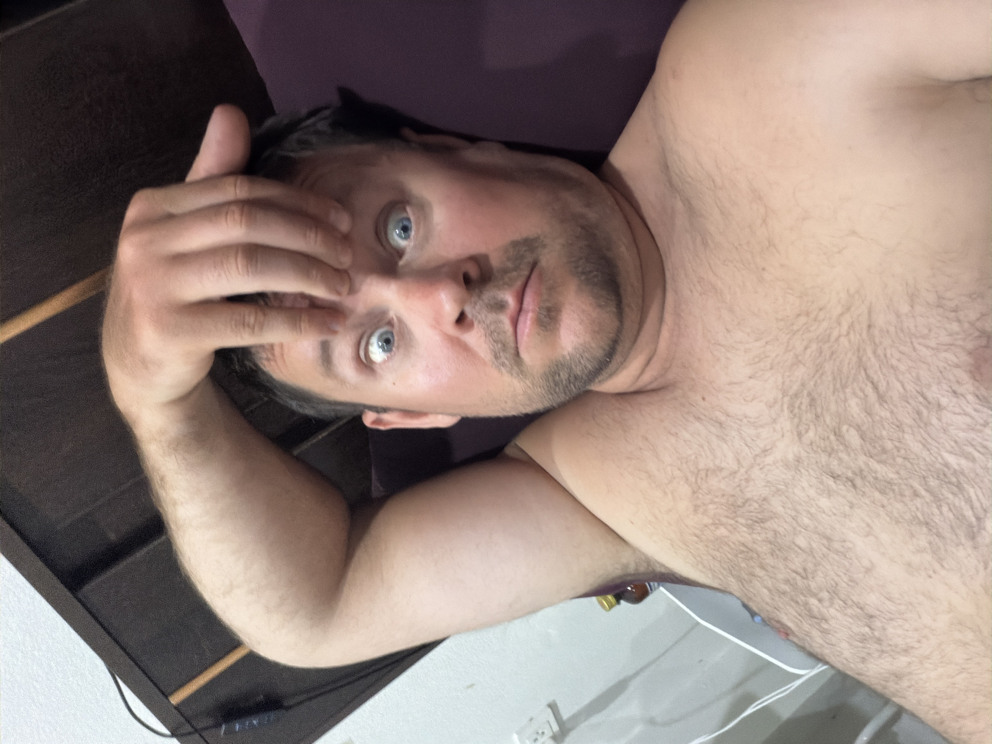 A man, appearing to be in his late 30s or early 40s, is captured in a selfie-style photograph taken in what is identified as Baan Mae Nam, Thailand. The man is lying down, possibly in bed, with his left arm bent and resting on his forehead. His eyes are wide open, giving him a surprised or startled expression. He has short brown hair and a light beard. He is shirtless, revealing a hairy chest and shoulder. In the background, a dark brown headboard is visible, along with a wall with a light switch. To the left of the man, a white electrical cord is plugged into the wall. A small brown bottle with a yellow cap is also visible near the man's arm. The overall environment suggests a personal living space, likely a bedroom.
