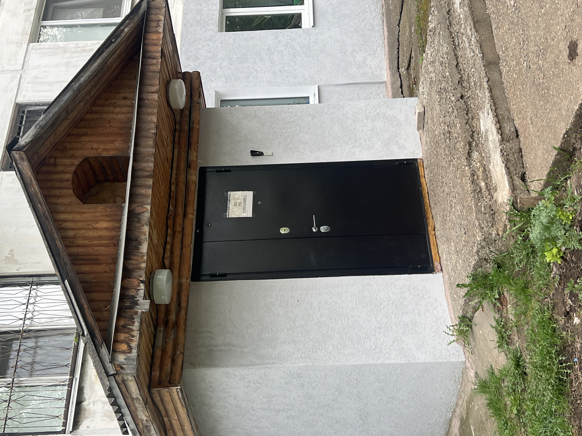 The image shows the entrance to a building in Chaikovsky, Russia. The entrance features a black metal door under a wooden A-frame porch. The building's exterior is light gray with a textured finish. There are two white-framed windows above the door. A sign is attached to the door. The sidewalk is cracked and overgrown with vegetation.