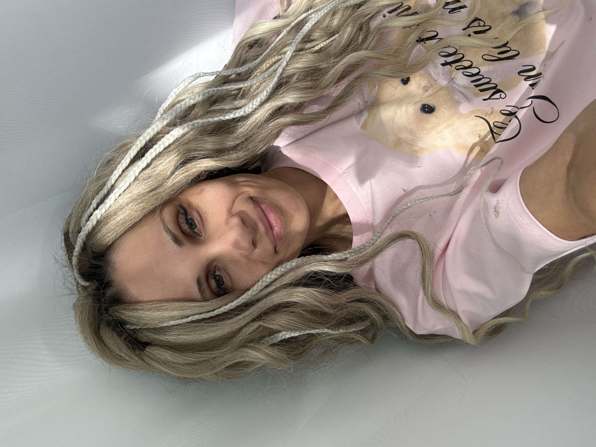 A woman with blonde and white braided hair is lying down, wearing a light pink pajama top with a teddy bear graphic and text that says "Sweetest...". The background is a plain white surface.