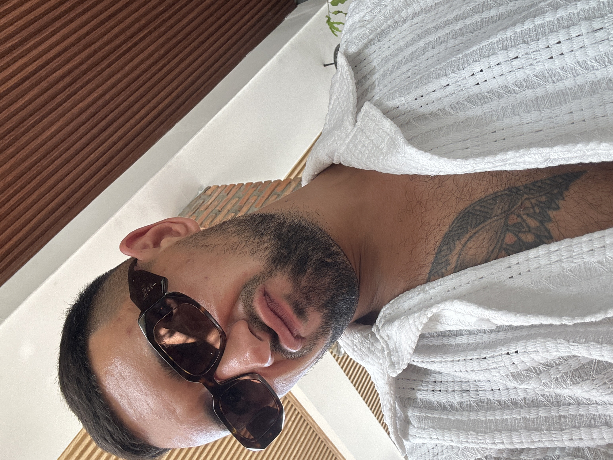 A man with a short haircut, a beard, and sunglasses makes a pouty face while looking into the camera. He is wearing a white open-collared shirt with a textured pattern. A portion of a tattoo is visible on his chest and shoulder. The background consists of wooden slats and a white architectural element. The overall impression is that the photograph was taken outdoors, possibly in a semi-private or resort-like setting in Nam Nha Trang Ward, Vietnam.