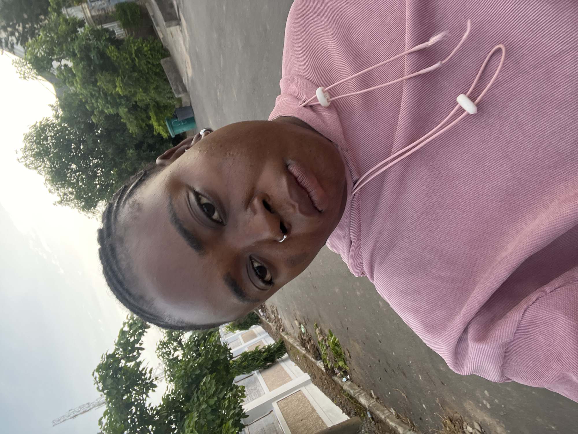 A selfie captures a person from the chest up, leaning slightly to their right, with the camera held at an upward angle. The person is wearing a pink hooded sweatshirt with drawstrings. Their hair is braided. They have a nose ring and an earring. The background shows a street scene in Abuja, Nigeria. To the left, a low, ornate wall lines the street, with some debris and vegetation at its base. Beyond the wall, trees and structures are visible. To the right, more trees and buildings are seen, along with two trash bins. The sky above is overcast, with the sun creating a bright, hazy light.