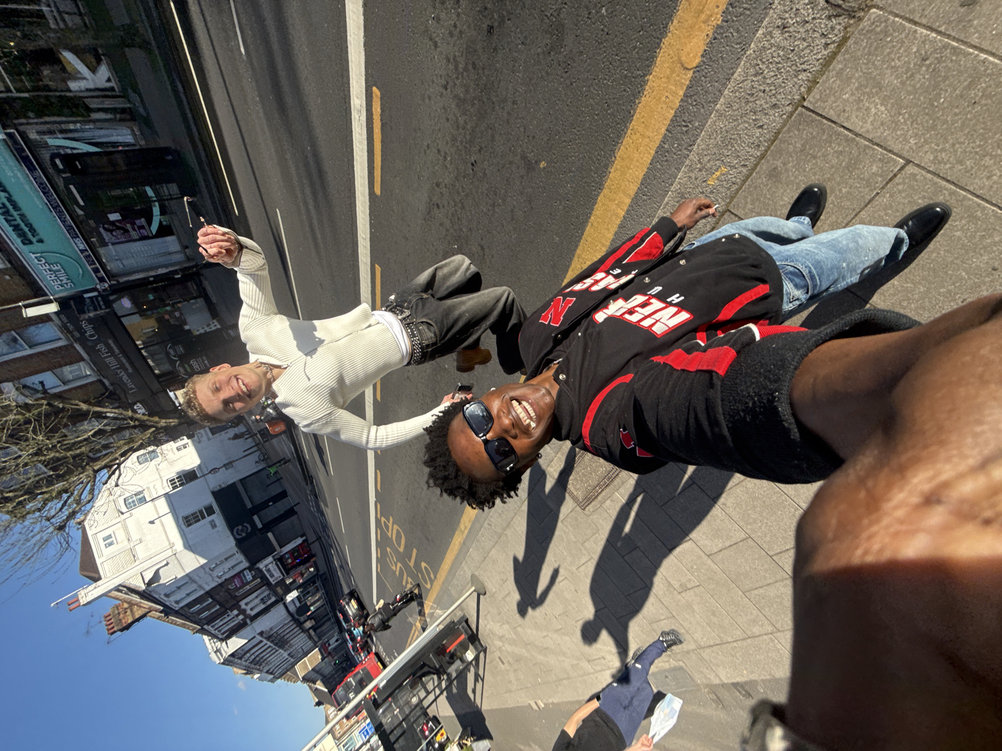 This is a low-angle photograph of two young men standing on a pavement next to a road. The man on the left is light-skinned, wearing a white ribbed outfit and has short blond hair. He is holding a cigarette in his right hand. The man on the right is dark-skinned, wearing a black and red jacket and sunglasses. He is smiling. They are standing next to a busy London street. Buildings, including shops and residential properties, are visible in the background. A red double-decker bus is also visible. Street signs and advertisement boards are mounted to the buildings, including an advertisement board for a fish and chip shop named "Lavender Hill Fish Chips". The sign also mentions “cash” and “New NHS patients welcome”. Yellow lines mark the edge of the road next to the sidewalk. The picture may have been taken on Lavender Hill Street in London, United Kingdom. The photo is taken in daylight and possibly taken with a wide angle lens. The view of the ground is distorted from a phone’s camera lens, causing it to curve.