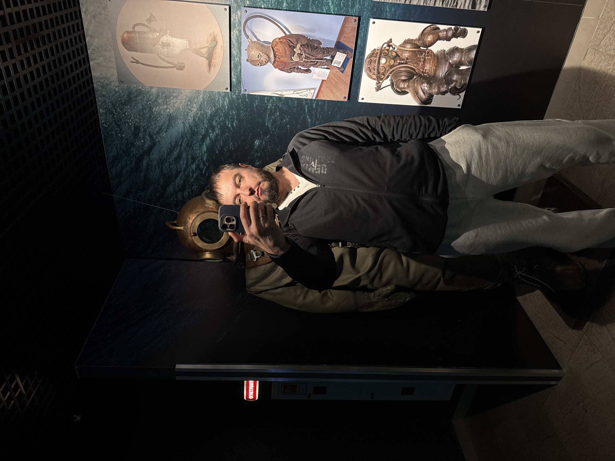 A man is captured in a selfie reflection, standing in what appears to be a museum or exhibition dedicated to diving. The image is taken in portrait orientation, showing the man from the waist up, holding a dark smartphone to take the picture. He has a beard, short dark hair, and wears a dark zip-up jacket over a light top and light grey sweatpants. A ring is visible on his left-hand ring finger.
In front of him, on a dark, reflective display surface, is a brass diving helmet with multiple circular windows. Behind the man, on a wall with a dark blue-green, textured pattern, are three framed images. From left to right, these images depict: an early, somewhat faded illustration of a diver; a brown, old-fashioned diving suit with a helmet and air hose; and an intricate, metallic, rigid diving suit or device.
The man's jacket has partially visible white text on the left chest area, which includes the word "TRAINING." Below the main display counter, a rectangular object has a red vertical label with the word "LUMITEXNO" written in white. The lighting is artificial, suggesting an indoor setting. The floor is light-colored, possibly wood or tile. The overall scene suggests a historical or technological display related to underwater exploration, located in Kogalym, Russia.