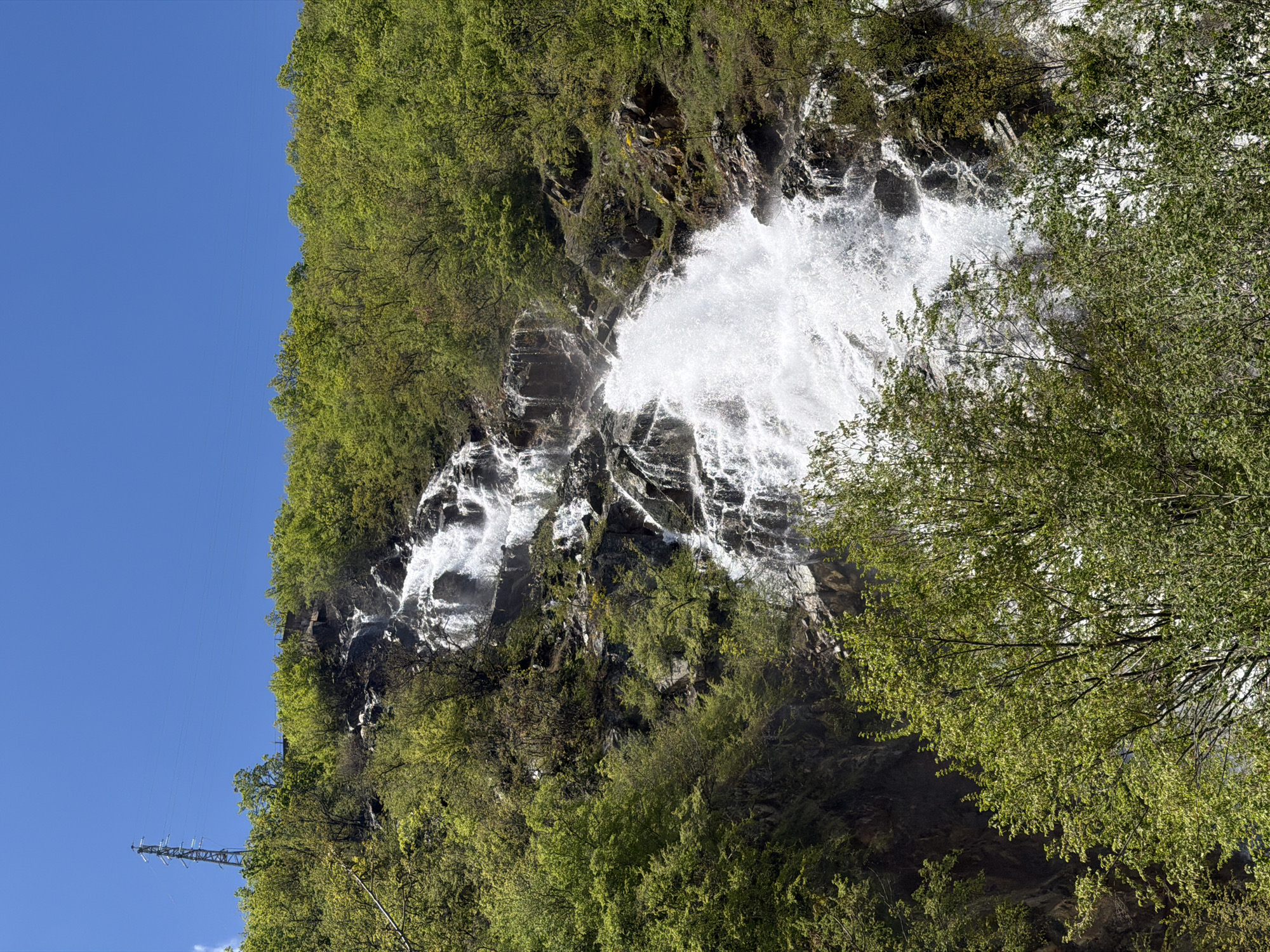 A waterfall plunges down a rocky cliff face covered in green trees, adjacent to a clear blue sky. The waterfall is the central feature of the image, with white water cascading over dark rocks. Green trees are present on the cliff face and in the foreground. On the left of the image, a section of clear blue sky is visible. In the bottom-left corner, a metal utility pole can be seen. The scene is in Pastra, Bulgaria.
