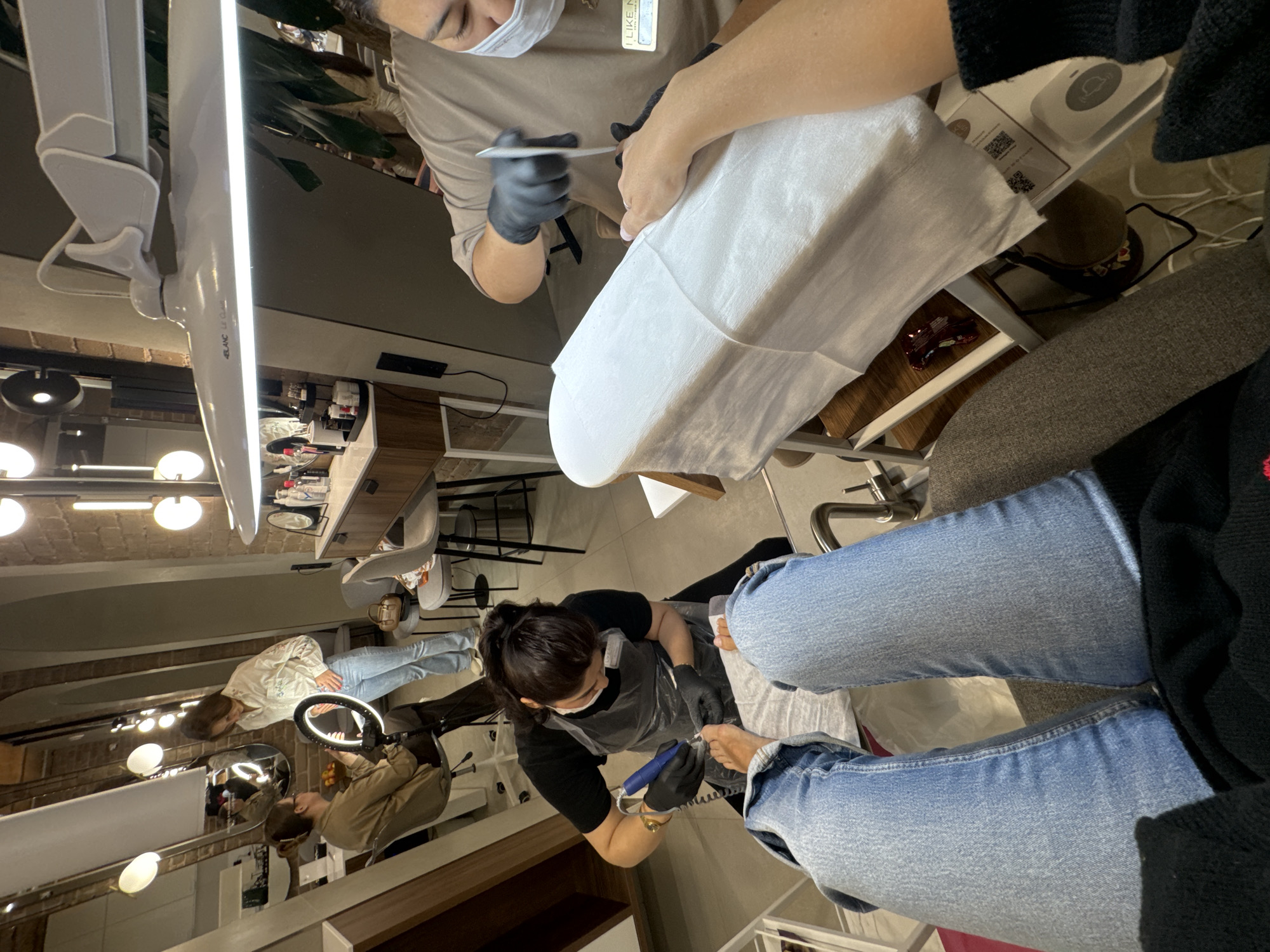A high-angle shot shows a pedicure and manicure being performed inside a beauty salon, presumably located in Moscow, Russia. There are two manicure tables, with a manicurist at each one, attending to clients. The clients are seated, and the manicurists are standing, wearing face masks and gloves for safety. One manicurist wears a light brown uniform top and the other wears a black top. The client's legs and hands can be seen with the manicurists working on the customer's nails. Professional lighting illuminates the work areas. Salon décor includes brickwork and modern lighting fixtures.