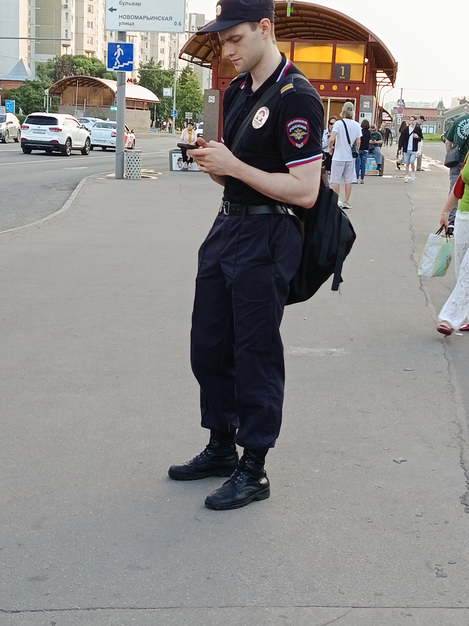In Moscow, Russia, a police officer stands on a sidewalk, looking down at his phone. He is wearing a dark blue uniform with a badge on his left sleeve. A black backpack is slung over his left shoulder. The sidewalk is paved and adjacent to a street with traffic, including white cars and a white SUV. In the background, there are buildings, a bus stop with a red roof, and other pedestrians. A sign indicating "Novomaryinskaya Street" is visible, with a distance of 0.6 km. The officer's feet are clad in black boots.