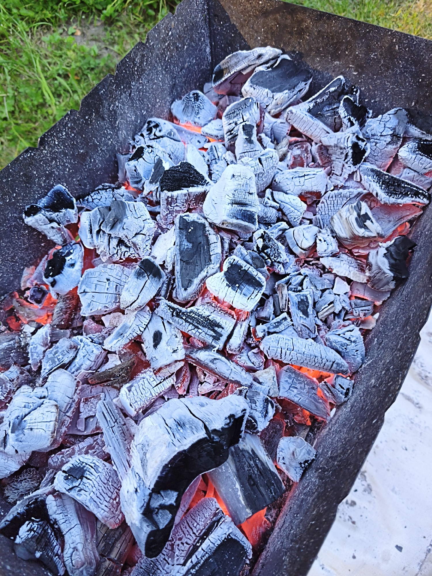 A close-up, overhead view shows glowing red embers and white-grey ash-covered charcoal pieces within a dark metal barbecue grill. The grill is placed on a light-colored surface, possibly concrete or a tarp, with green grass visible in the upper left corner, suggesting an outdoor setting. The scene is devoid of any people. The lighting is natural, with the bright glow of the coals illuminating the surrounding charcoal fragments. The image appears to be taken in Maardu, Estonia, based on the provided context.
