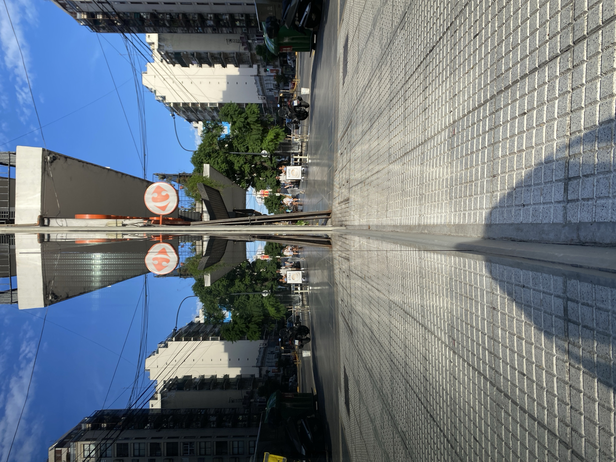 The image depicts a street scene in Buenos Aires, Argentina, seemingly reflected in a polished surface or puddle, giving it a mirrored effect. Buildings, a traffic/business sign with a smiley face, trees, and pedestrian activity are visible. The right side of the image shows textured pavement in a running bond pattern. A few people are walking in the distance. There are parked vehicles along the street and power lines above. The sky is blue with clouds.