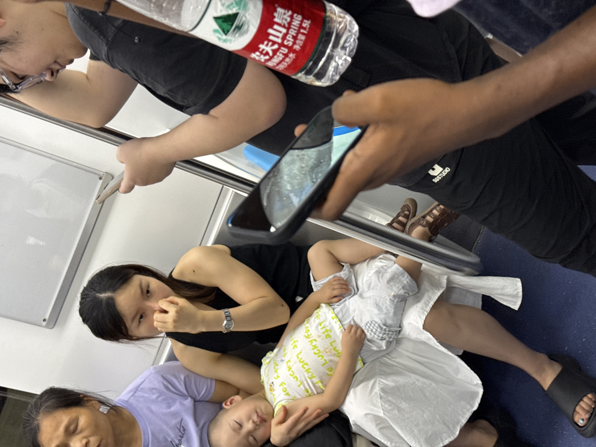 In an interior setting that appears to be a subway car, a young child sleeps on the lap of an adult woman. The child is wearing a white t-shirt with yellow lettering and light-colored shorts. The woman, wearing a black sleeveless top and a watch on her left wrist, has her hand near her face. To the left, another woman in a purple t-shirt is visible. In the foreground, a person holds a smartphone, its screen illuminated, and a bottle of Nongfu Spring water is positioned to the right. Other individuals are partially visible, with one person in a black t-shirt holding onto a vertical pole. The environment is characterized by grey and white interior walls and a blue floor.