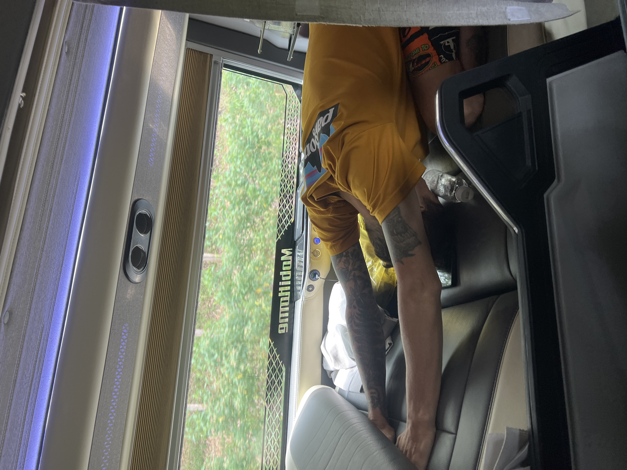 An interior view shows a person bending over in what appears to be a passenger seat of a bus or large van. The person wears a yellow t-shirt with a "Patagonia" logo and has tattoos on their arms. The seats are upholstered in a light gray and dark gray material with white accents. A black panel with a metallic trim is visible to the right of the seat, and a window on the left shows green trees and vegetation outside. Part of the word "Kiah" is seen reversed on the window frame. A water bottle is visible near the person. The setting implies a vehicle traveling through Kon Ra Kiah, Vietnam.