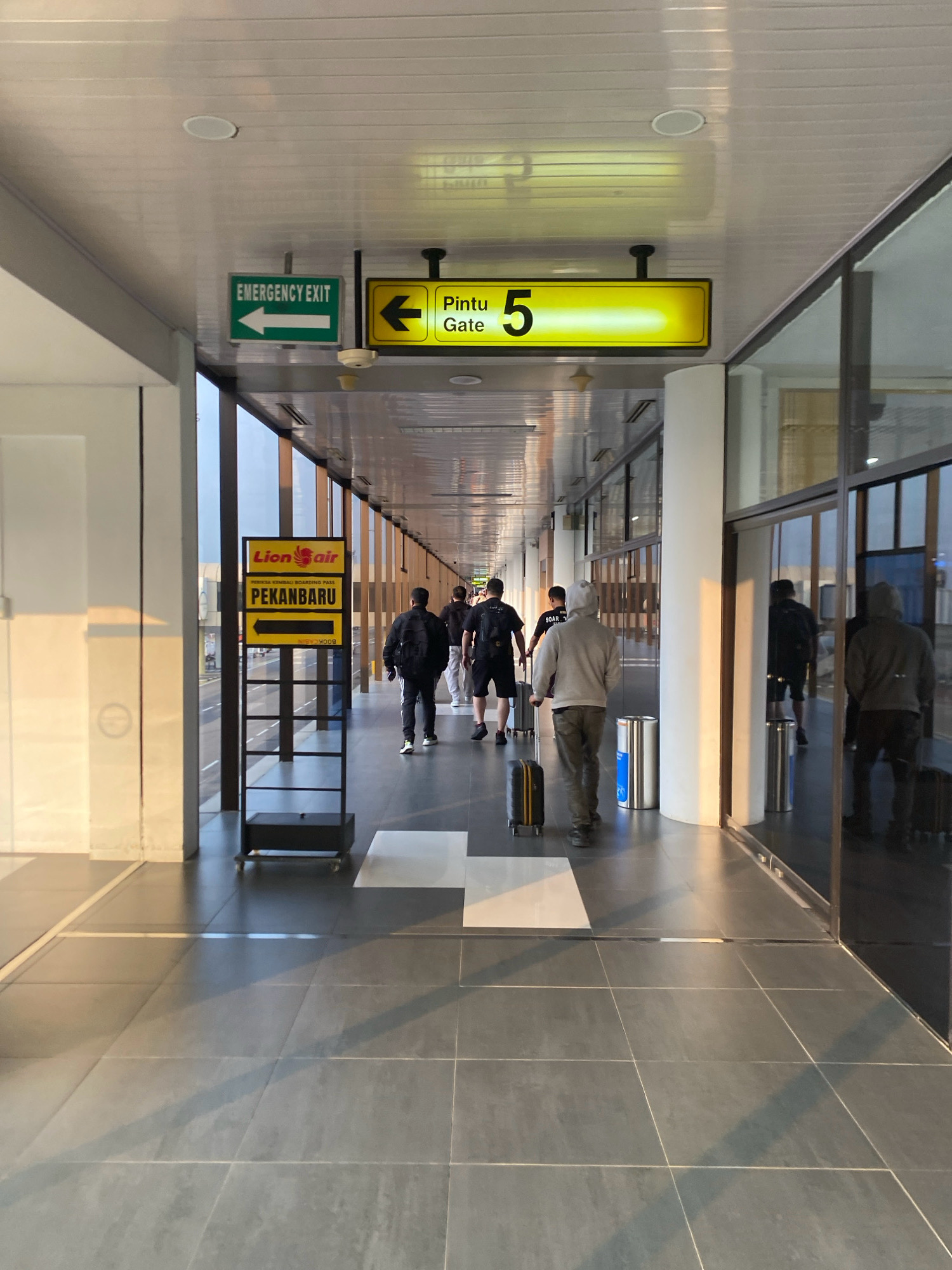 The image depicts an interior view of an airport walkway in Batam, Indonesia. Several individuals are walking down the corridor, some with luggage, suggesting they are passengers. Overhead signs indicate "EMERGENCY EXIT" with an arrow pointing left and "Pintu Gate 5" with an arrow pointing left. A sign for "Lion Air PEKANBARU" is also visible, further confirming an airport setting and a destination within Indonesia. The environment is modern with a tiled floor, white ceiling, and large glass windows lining one side of the walkway, offering a view of the tarmac or an outdoor area. The lighting suggests it is either early morning or late afternoon, given the warm sunlight casting shadows.