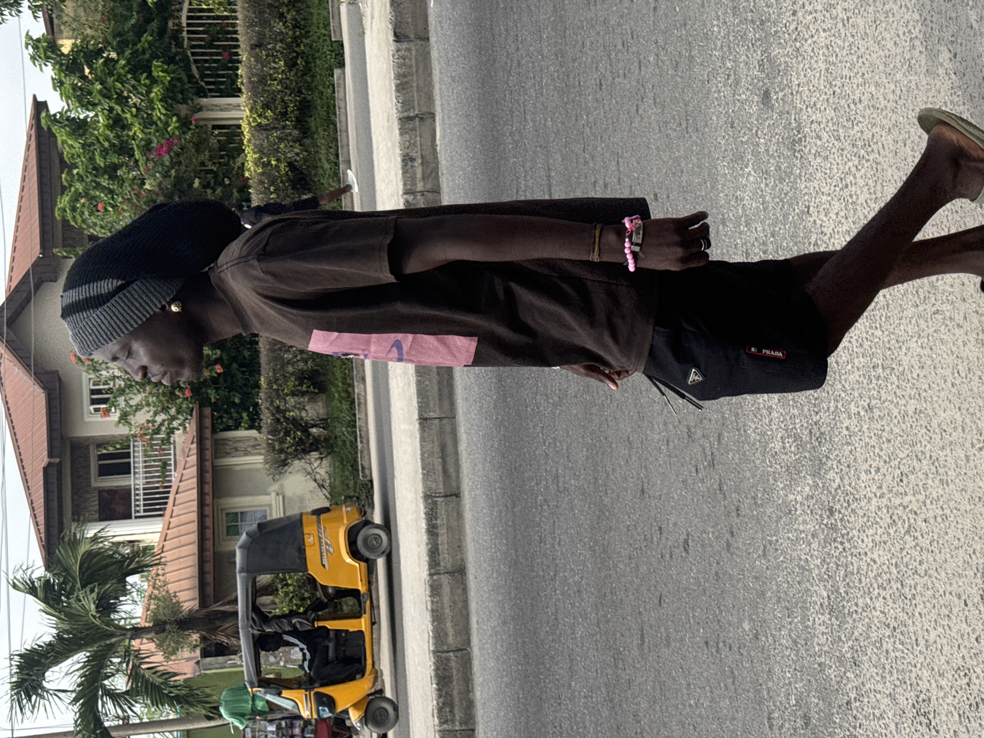 An outdoor, eye-level shot, originally rotated 90 degrees counter-clockwise, depicts a person standing by a road in what appears to be Ado, Nigeria. The subject, a person with dark skin and a neutral expression, occupies the right side of the frame, facing left. They wear a black beanie with grey stripes, a dark brown or black short-sleeved t-shirt with an indistinct pink graphic on the chest, and black shorts. Their feet are clad in light-colored flip-flops. Visible accessories include a gold-colored earring, a pink beaded bracelet, and a brown cord bracelet on their right wrist. The shorts feature a white rectangular label with "PRADA" written in black text on the left leg.
In the mid-ground, a yellow motorized tricycle, commonly known as a keke napep, is parked on the side of the road, partially obscured by lush green foliage and a tall palm tree. A person is visible sitting inside the keke. Behind the vegetation and keke napep, a multi-story building with a reddish-brown roof and white balconies can be seen. The setting includes an asphalt road, a grey concrete curb, and a sidewalk. The time of day appears to be daytime, with clear, bright weather. No other specific text is clearly legible on any other objects or signs within the image.