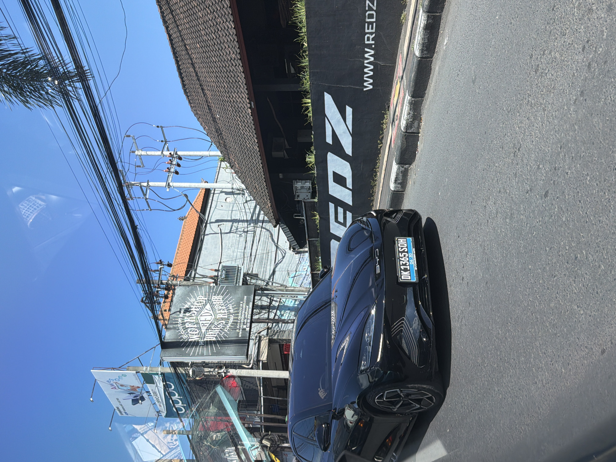 The image captures a vibrant street scene in North Kuta, Indonesia, on a clear, sunny day. The perspective is from inside a vehicle, looking towards the right side of the road.
In the foreground, a sleek black car, likely a BYD based on a visible logo, is prominently featured. Its front license plate, "DK 1365 SQH," with "06-30" below, indicates its registration in Bali. The car's modern design is highlighted, with intricate patterns on its front bumper/fender area.
The background reveals a bustling urban environment. To the right, a black building features large white lettering spelling "REDZ" and "WWW.REDZ," alongside "WWW.REDZSURFBOARD.COM," suggesting a surf-related business. This building has a distinctive dark, tiled roof. Further back and to the left, another building with a reddish-brown tiled roof is visible. A dense network of overhead power lines and utility poles crisscrosses the clear blue sky.
Several signs and billboards are scattered throughout the scene. A large black sign with a diamond-shaped logo partially reads "REDZ SURFBOARDS & REPAIRS," and includes an address "JL. RAYA CANGGU 18 X" and a phone number "+62 81 87". Other advertising signs display brand names like "OPPO A98," "oddo," and "SAMSUNG," with some partially obscured text like "Awe M" and "GASTADSG." A palm tree's fronds are visible on the far left. The road surface is asphalt, and a painted curb, featuring black, white, and yellow sections, marks the edge of the street.