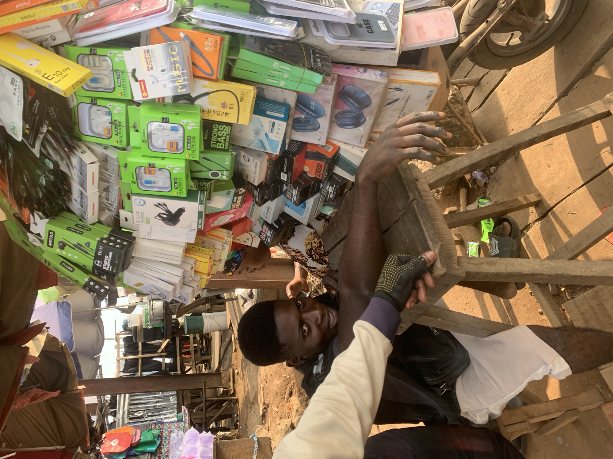 The image depicts an outdoor market stall in Shagamu, Nigeria, selling various electronic accessories, primarily mobile phone accessories like earphones and chargers. In the foreground, a young Black man, dressed in a black t-shirt, white shorts, and wearing a black fanny pack, is seated behind a wooden stall. He is looking towards the camera with a slight smile and his left hand is resting on the stall. Another person's arm and hand, wearing a light-colored sleeve and a black glove with yellow dots, is visible in the lower left corner, appearing to interact with the seated man or the stall. The stall is laden with colorful packaging of electronic items, including headphones, data cables, and phone cases, arranged on a display rack. In the background, other market stalls, wooden structures, and a motorcycle wheel are visible, suggesting a bustling market environment. The ground is made of wooden planks, and the scene is illuminated by natural sunlight.