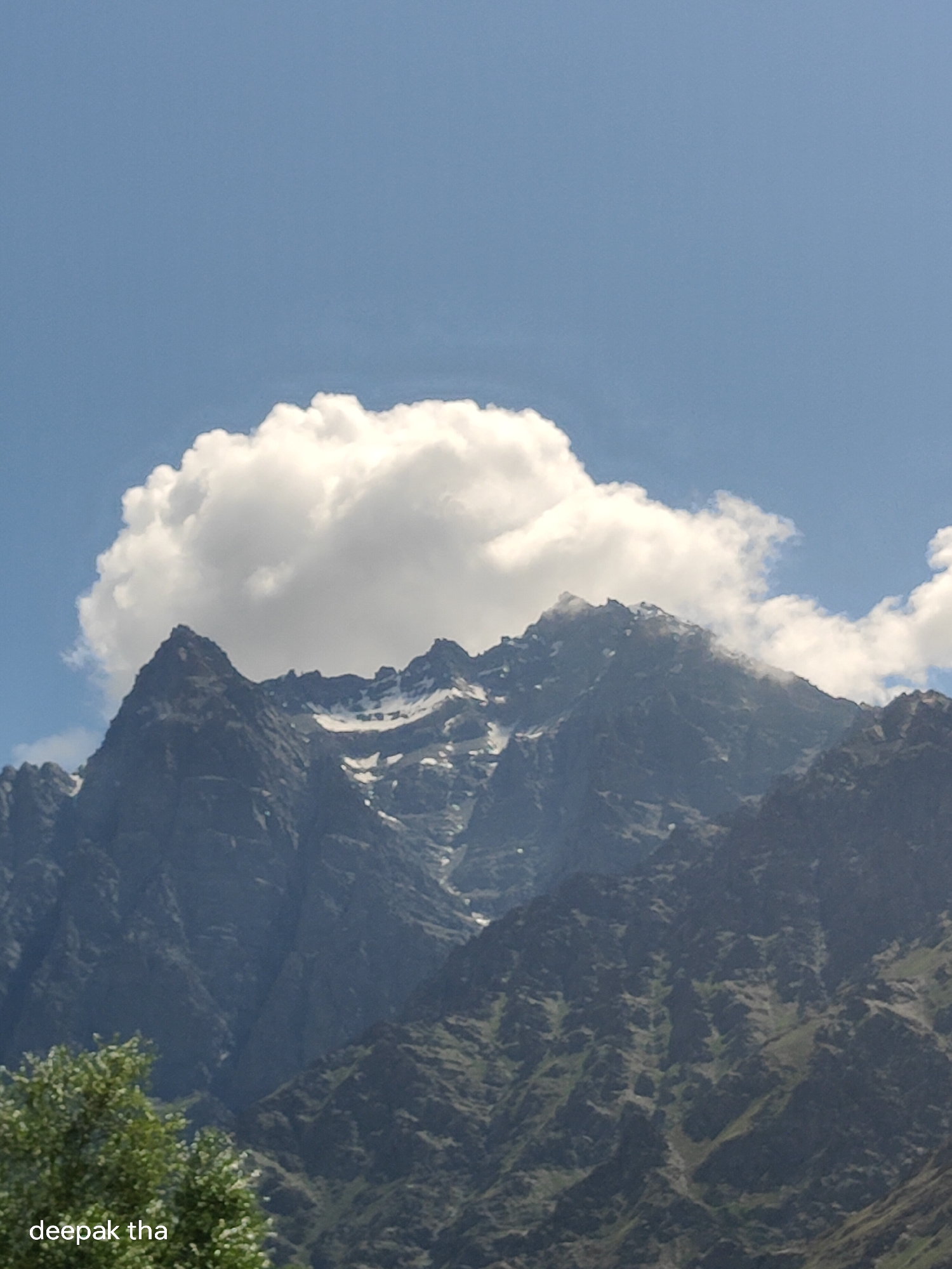 A large, fluffy white cloud floats above a jagged, dark mountain range under a clear blue sky. Patches of snow dot the upper reaches of the mountains. In the foreground, lush green trees are visible on the left side of the frame, partially obscuring the lower slopes of the mountains. The text "deepak tha" is superimposed on the bottom left corner of the image. The scene appears to be a natural landscape in Darcha, India.