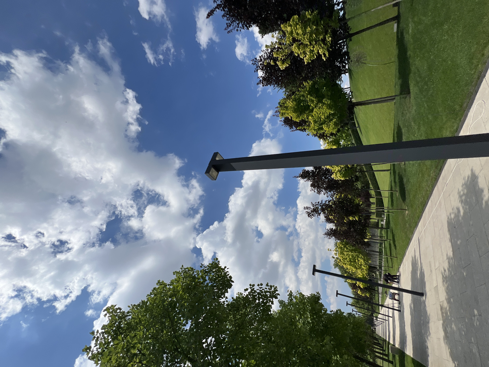 The media file depicts an outdoor scene in a park-like setting, with a paved walkway stretching into the distance. The walkway is lined with trees on one side and a neatly manicured lawn on the other. Several lampposts are visible along the walkway, casting shadows on the pavement. In the foreground, a large lamppost with a modern design dominates the frame. A few people are scattered along the walkway, some walking and others appearing to be socializing near the lampposts. The sky is a vibrant blue with scattered white clouds, suggesting a pleasant day. The overall atmosphere appears to be peaceful and relaxing, indicative of a leisurely stroll in a well-maintained public space. The location cues, such as the distinct style of the lampposts and the type of trees, along with the context provided, suggest Krasnodar, Russia. The time of day appears to be daytime, given the lighting and shadows. There is no visible text in the image.
