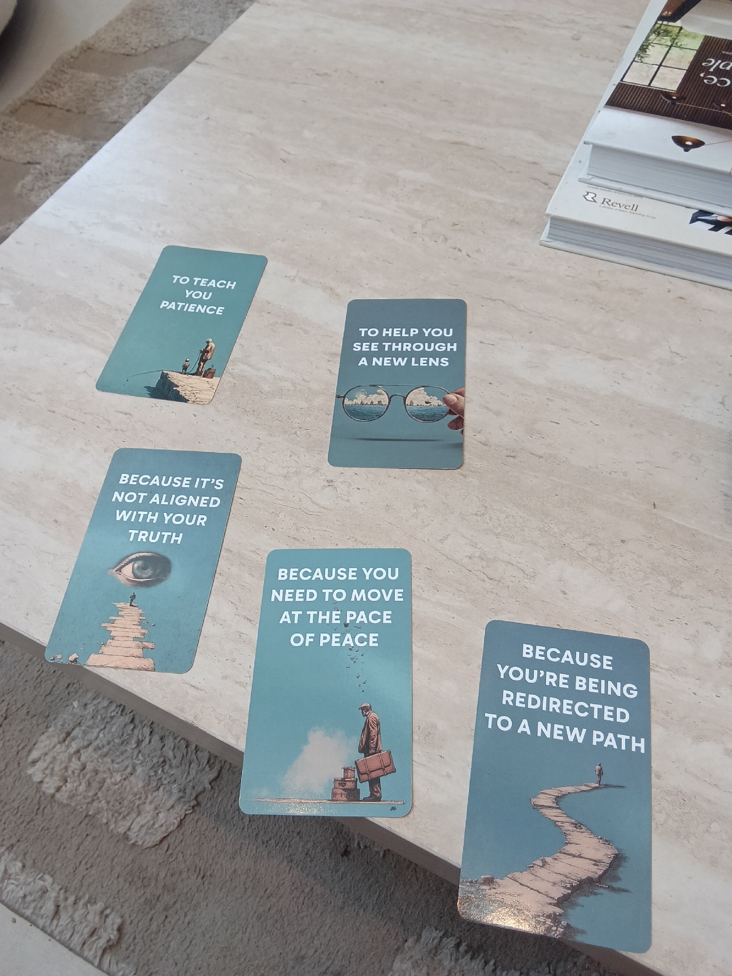 The image shows five teal-colored cards with white text and surreal illustrations, arranged on a light-colored, patterned surface that suggests a marble or similar material. To the right of the cards is a stack of books or magazines, the topmost bearing the word "Revell." The cards contain phrases: "TO TEACH YOU PATIENCE," depicting a small figure fishing with an adult; "TO HELP YOU SEE THROUGH A NEW LENS," showing hands holding eyeglasses; "BECAUSE IT'S NOT ALIGNED WITH YOUR TRUTH," illustrated with a path leading to an eye; "BECAUSE YOU NEED TO MOVE AT THE PACE OF PEACE," showing a figure with luggage; and "BECAUSE YOU'RE BEING REDIRECTED TO A NEW PATH," depicting a winding path with a figure at the end. The cards appear to be part of a deck designed for introspection or guidance. The location is not discernible from the image.