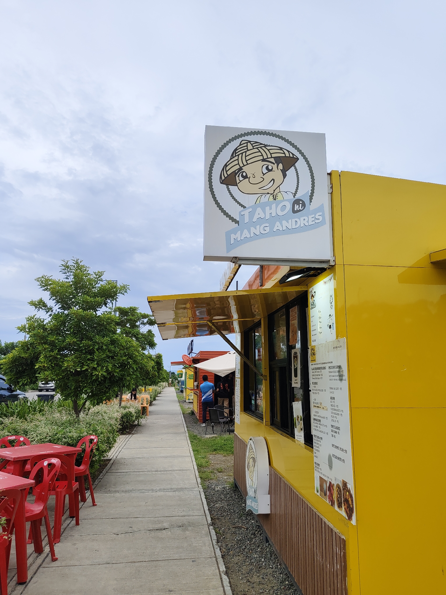This image shows a bright yellow food kiosk named "TAHO HI MANG ANDRES," located on a sidewalk in Davao City, Philippines. The kiosk has a sign featuring a cartoon character wearing a conical hat. A menu board is attached to the side of the kiosk. In the foreground, red tables and chairs are set up on the sidewalk, suggesting an outdoor dining area. A person wearing a blue shirt is seen standing at the counter of the kiosk. Beyond the kiosk, a row of trees and bushes line the sidewalk, with parked cars visible in the background. The sky is overcast with clouds. The scene depicts a casual commercial setting typical of many street-side food vendors in urban areas.