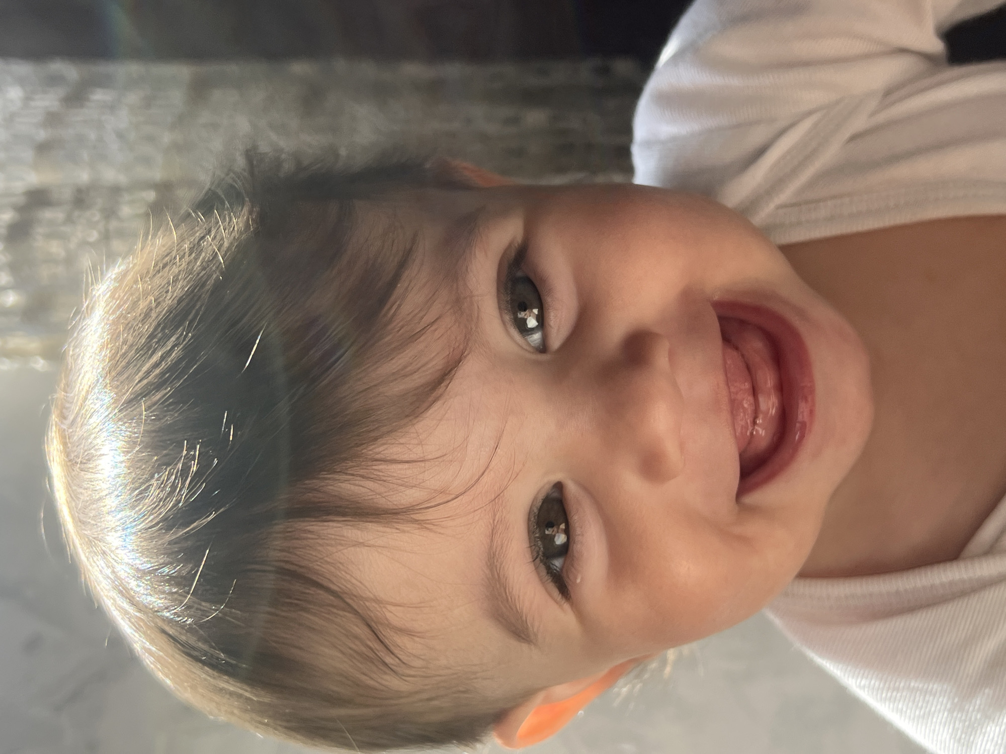 A close-up portrait shows a joyful baby, appearing to be a young toddler, positioned with their head to the left and body extending to the right due to the image being rotated 90 degrees clockwise. The baby is looking directly at the camera with a wide, open-mouthed smile, revealing a glimpse of their tongue.
The subject has dark brown hair, which is catching bright light, possibly sunlight, creating sparkling highlights on individual strands. Their large, dark eyes reflect the bright light source and possibly the surroundings or person taking the photo. The baby's skin tone is fair. They are wearing a simple white long-sleeved shirt or onesie. A small portion of their ear is visible.
The background is softly out of focus, suggesting an indoor setting with some indistinct textured surfaces behind the baby, possibly a wall or furniture. The strong light coming from the left illuminates the baby's face and hair, contributing to a bright and cheerful atmosphere. No specific location cues are visible within the image to confirm Elena, Bulgaria, but this is the provided context for the media file. There is no visible text. The overall mood conveyed is one of happiness and playfulness.