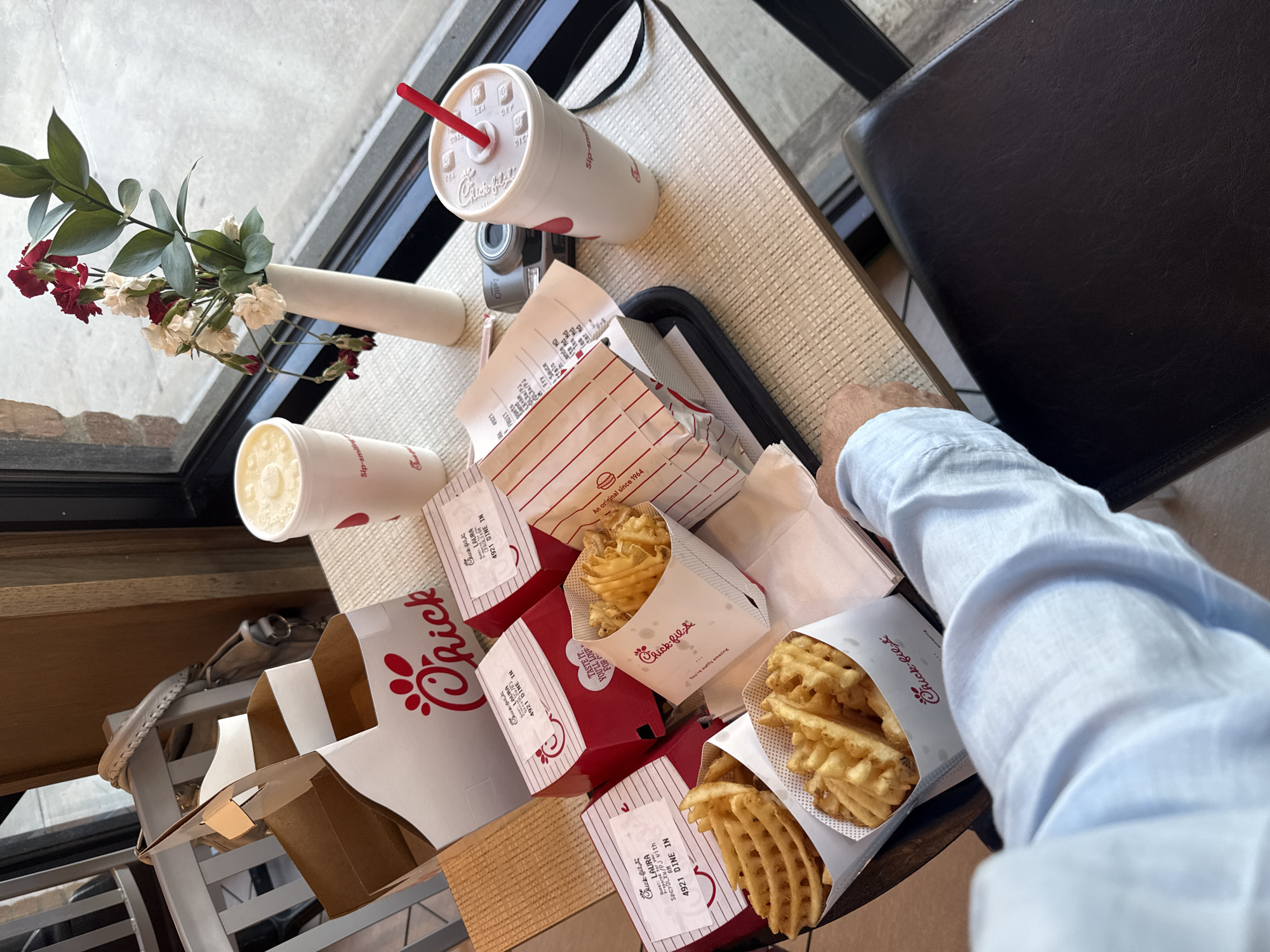 Here's a concise description of the image:
A high-angle shot depicts a meal from Chick-fil-A, including waffle fries, sandwiches, and drinks, arranged on a black serving tray on a light brown woven table. The presence of the name "LAURA 4921 DINE IN" on several items suggests the location is the Chick-fil-A at 4921 West Southlake Boulevard, Southlake, Texas, United States of America. A person wearing a light blue long-sleeved shirt is partially visible in the lower right corner, placing their hand near the tray. A small vase with artificial flowers sits next to the window in the background, near the camera and the drink. A digital camera also sits on the table next to the window. A black chair is visible in the background, suggesting this is an indoor restaurant setting.