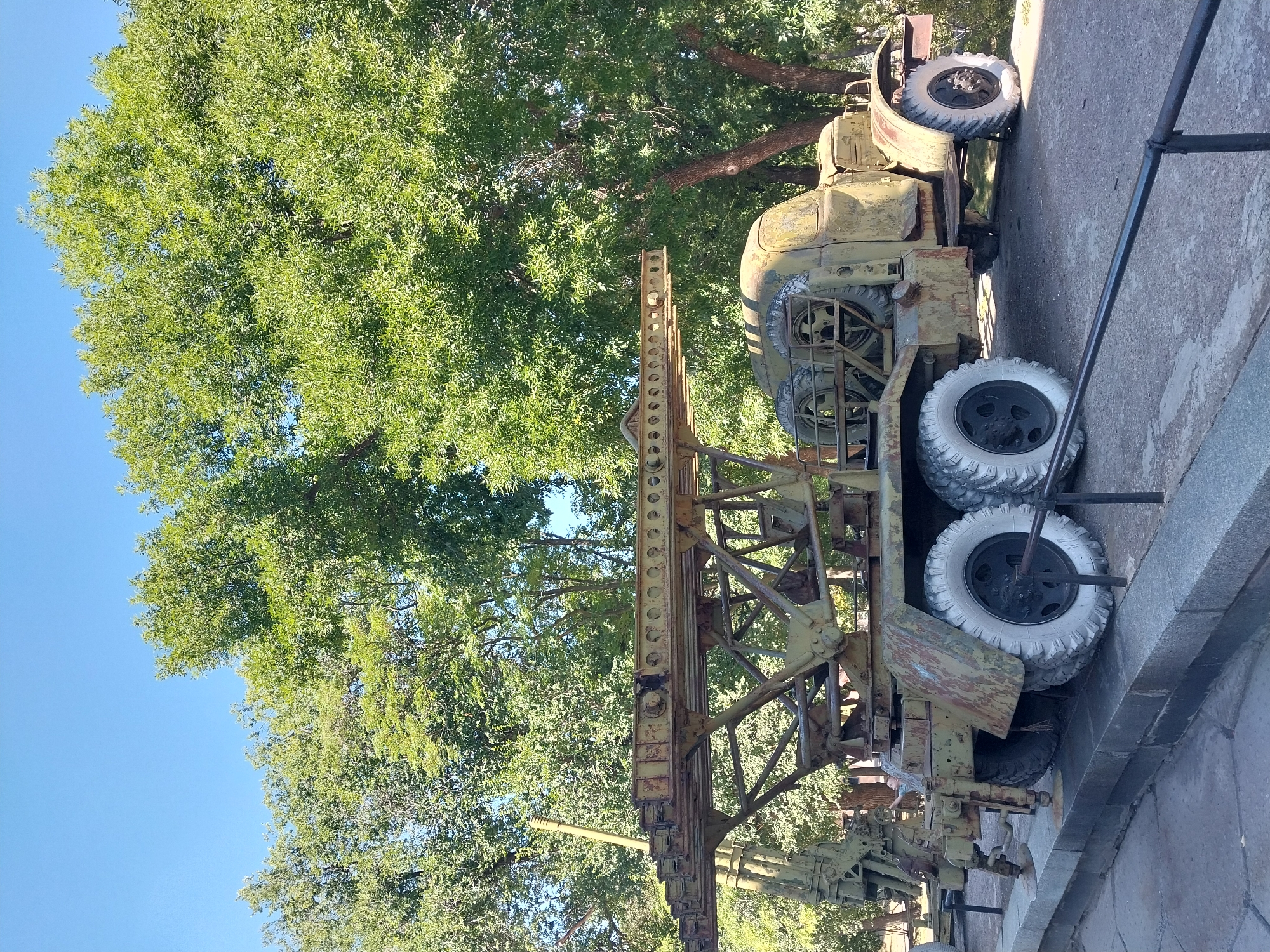 A weathered, olive-drab military vehicle, likely a rocket launcher on a truck chassis, sits outdoors in a park setting with lush green trees and a clear blue sky. The vehicle is positioned on a stone walkway with a black metal railing in the foreground. The truck has large, white-rimmed tires and appears to be old, with visible rust and wear. A person is visible in the background, near the front of the vehicle, but their features are not clear. The environment suggests a sunny day. This vehicle is located in the Alley of Heroes in the city of Dnipro, Ukraine, which is a memorial park featuring military equipment.