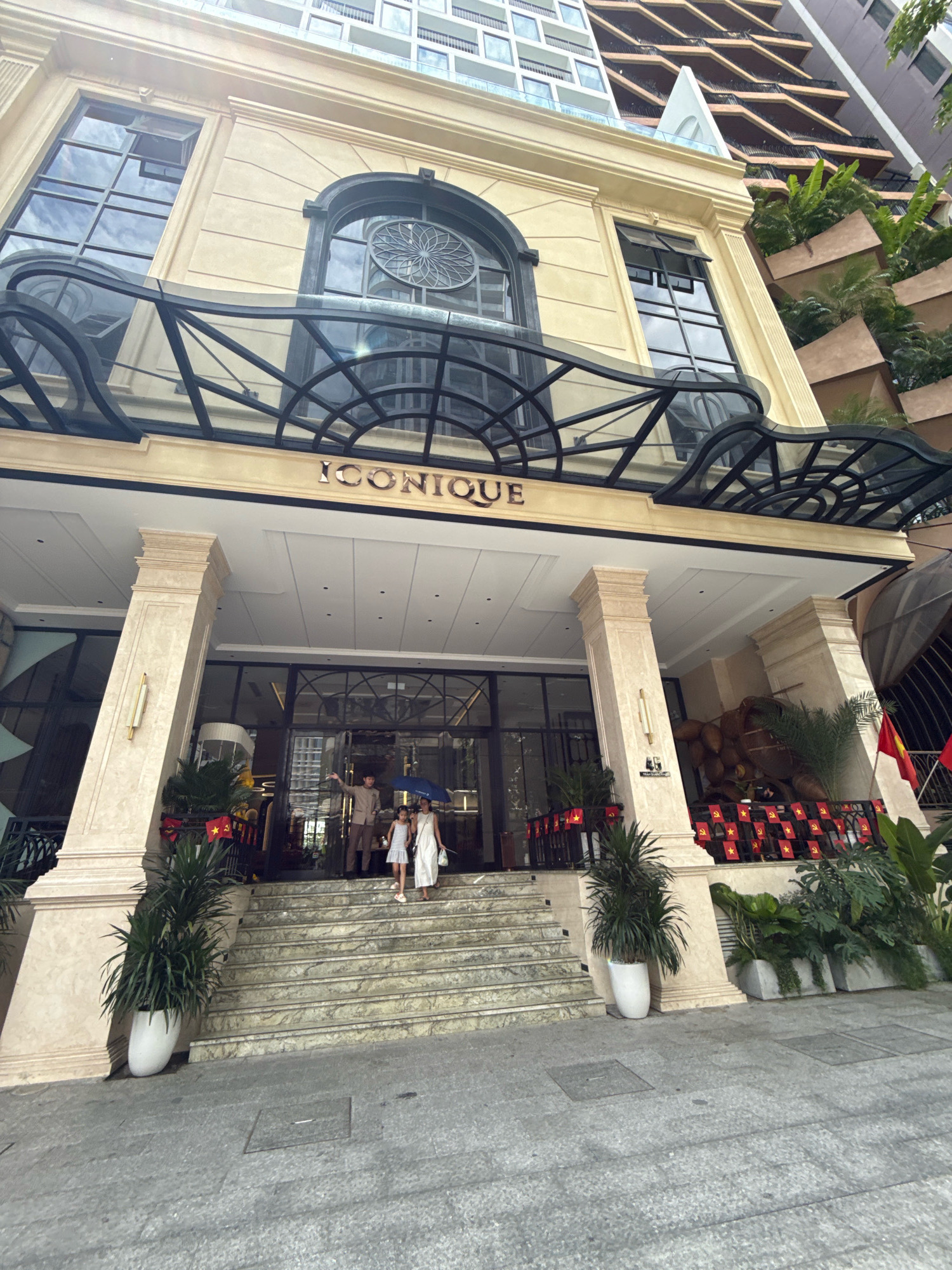 The image shows the exterior of a building with a sign that reads "ICONIQUE". The building is light yellow and has large windows. There are large pillars at the entrance, and a set of stairs leading up to the building. Two adults and one child are walking up the stairs. One of the adults is holding an umbrella. The building appears to be located in a city with other tall buildings in the background. The overall impression is that of a hotel or a luxury establishment. The flags visible on the building are likely Vietnamese flags, indicating the location is in Vietnam. Given the context of the flags and the general architectural style, the location is likely Nha Trang, Vietnam.